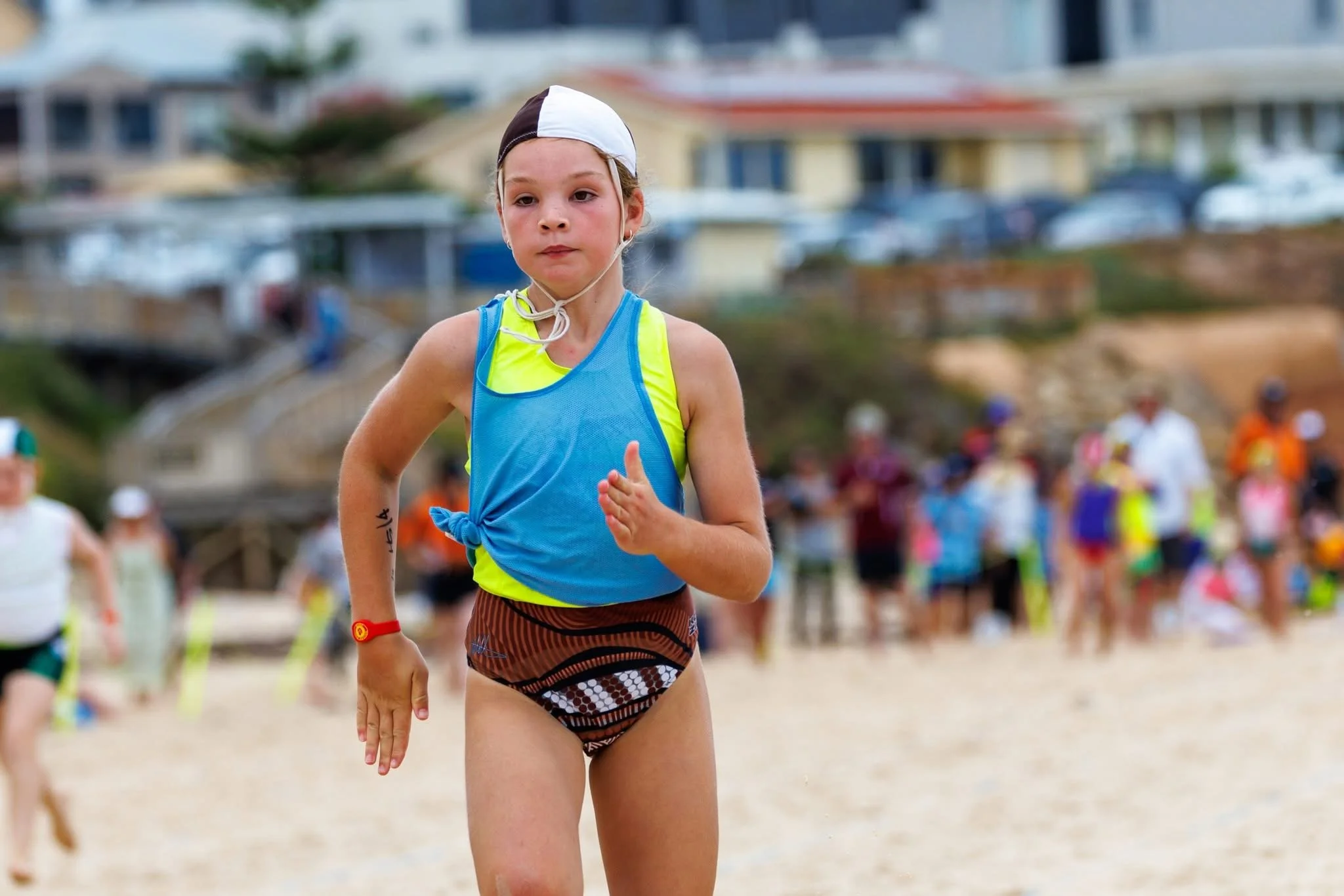 A young girl running on the beach during a race, wearing a blue tank top, a black and brown swimsuit, and a white and black swim cap, with a crowd of people in the background.