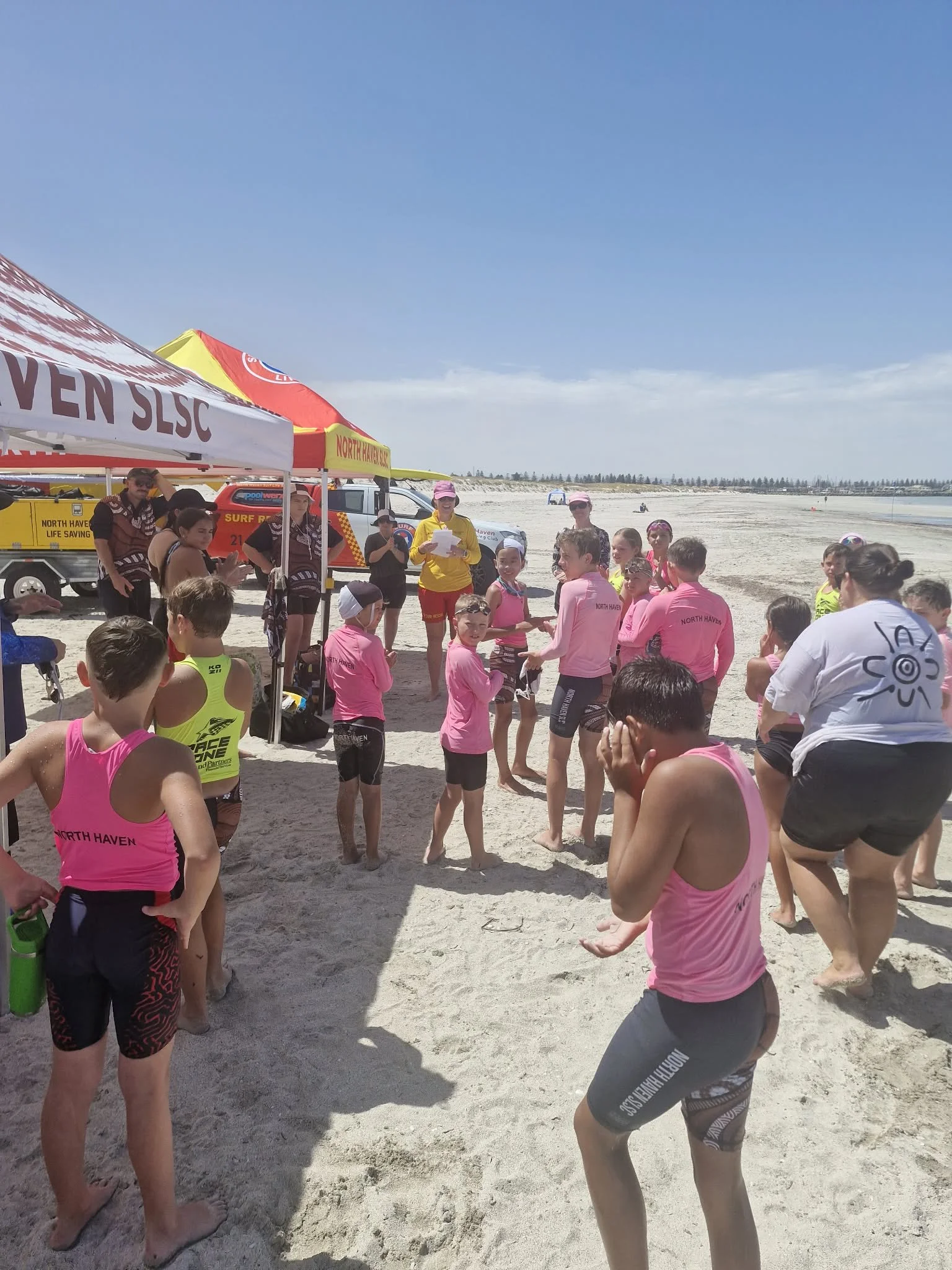Children and adults at a beach during a Lifesaving Club event, with tents, vehicles, and a clear blue sky in the background.