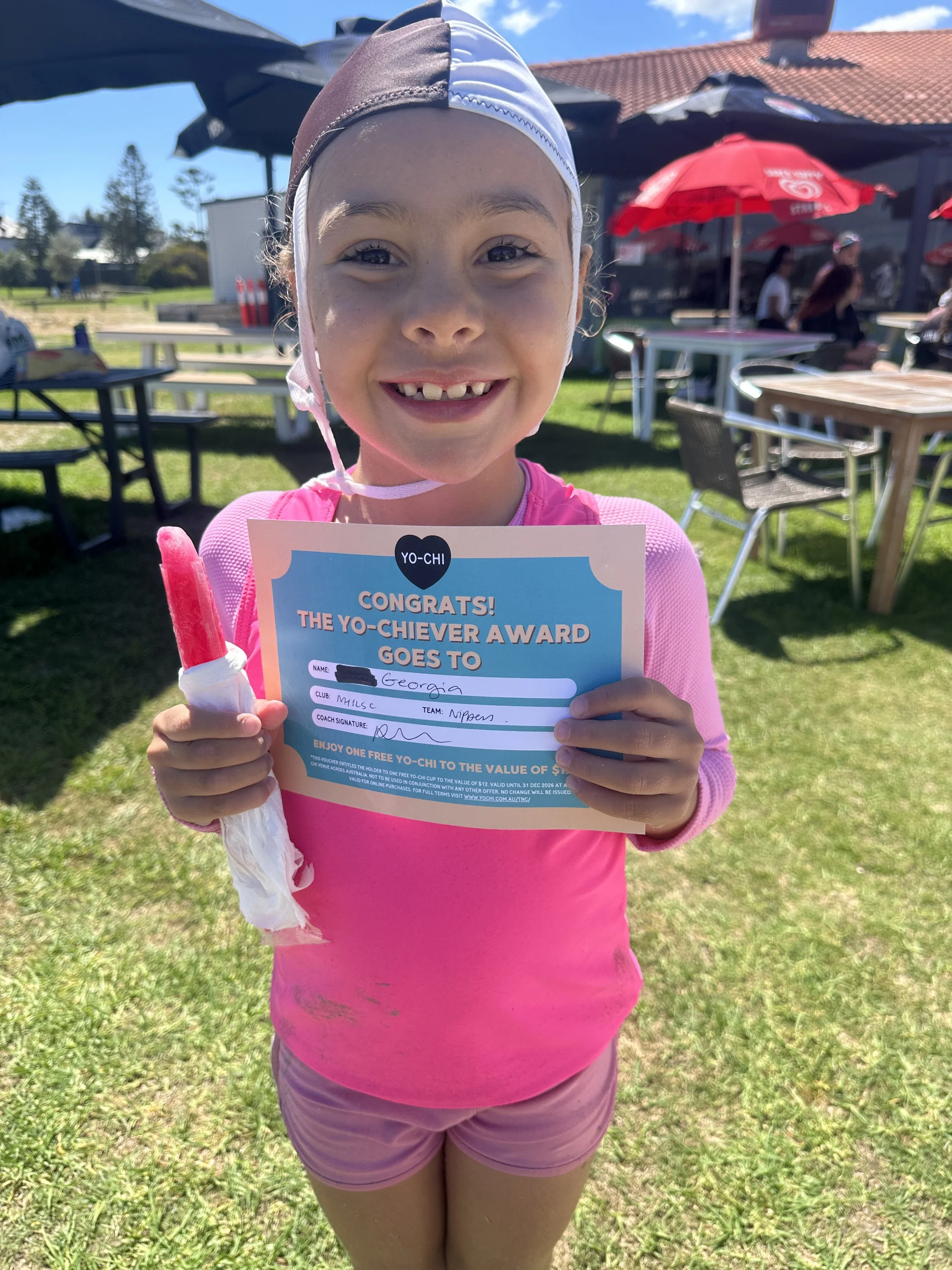 A young girl in a pink outfit holding a certificate and a popsicle, smiling outdoors on a sunny day with picnic tables and umbrellas in the background.