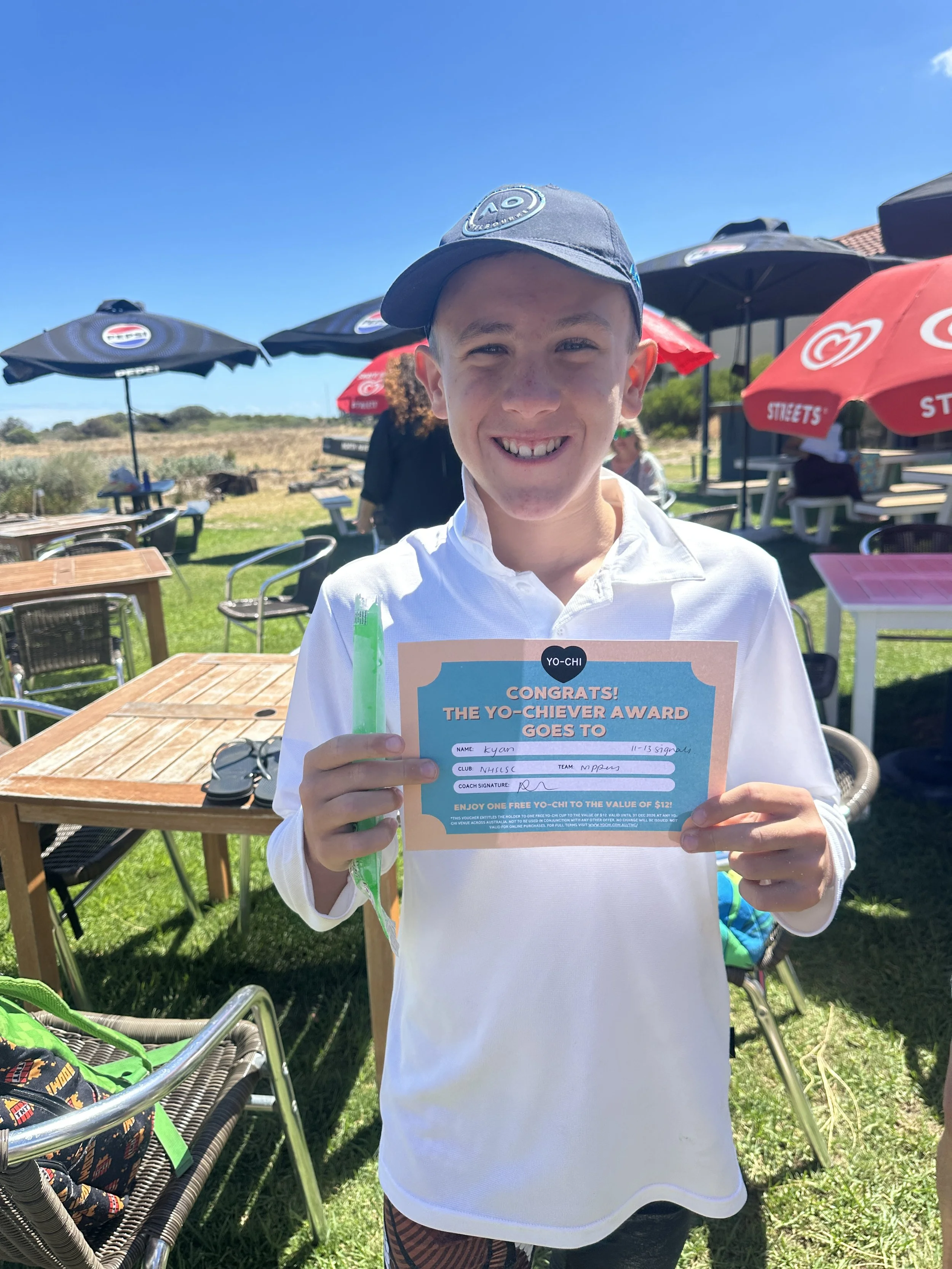 A young boy smiling outdoors at a sunny day, holding a certificate with the words "CONGRATS! THE YO-CHIAVER AWARD GOES TO," with a green pen in his right hand, standing among outdoor tables and umbrellas.