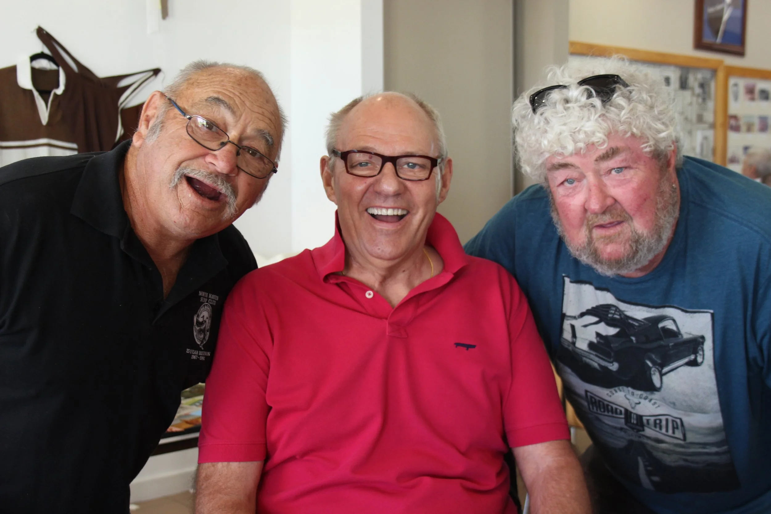 Three elderly men smiling and posing indoors. The man in the middle is wearing a red polo shirt and glasses, flanked by two men with longer gray or curly hair, one in a black shirt and the other in a blue shirt with a graphic design.