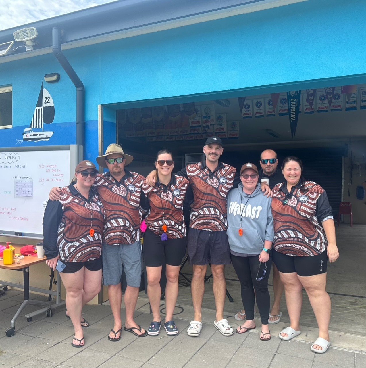 Group of seven people standing together at a beach event, wearing matching patterned shirts, some with sunglasses and hats, smiling, with signs and banners in the background.