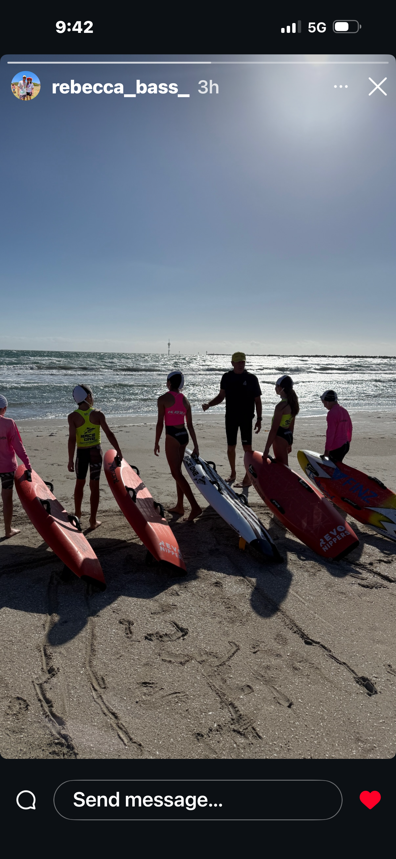 Group of young girls and an instructor preparing for a paddleboarding lesson on the beach, with paddleboards lined up on the sand and the ocean in the background under a clear sky.