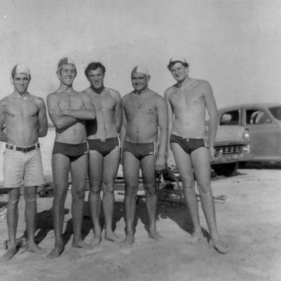 Black and white photo of five men in vintage swimwear and swim caps, standing on a beach, with a car in the background.