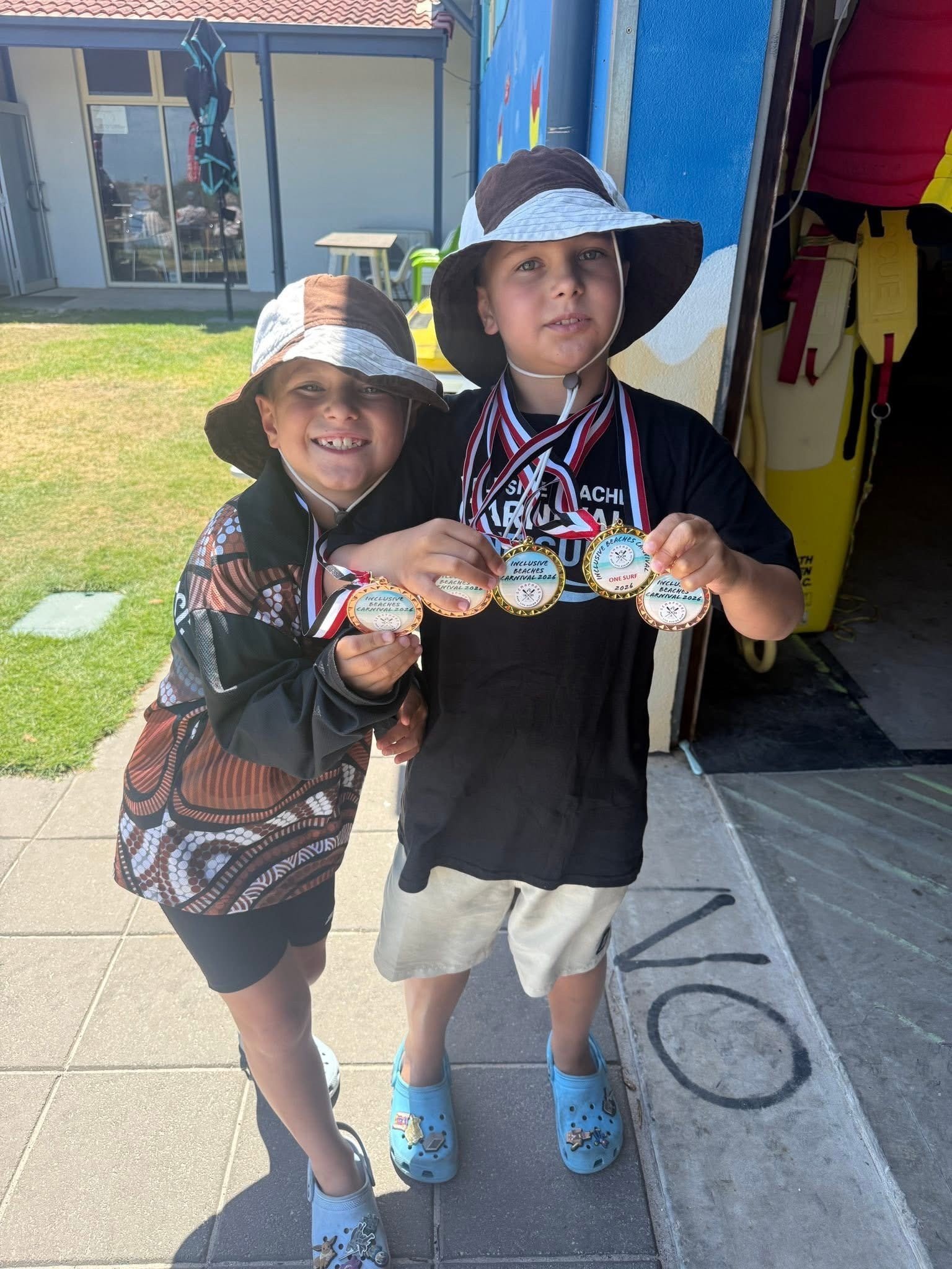 Two young children wearing hats and casual clothes, smiling and holding up medals they received from a beach or swimming event.