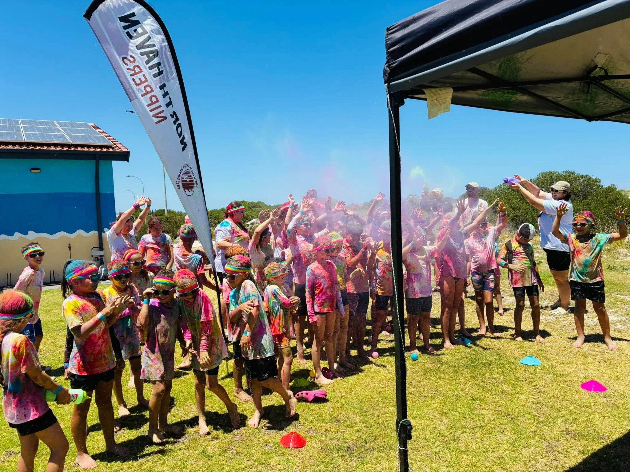 Children participating in a color powder festival outdoors, covered in colorful powder, with a woman spraying color on them, under a blue sky.