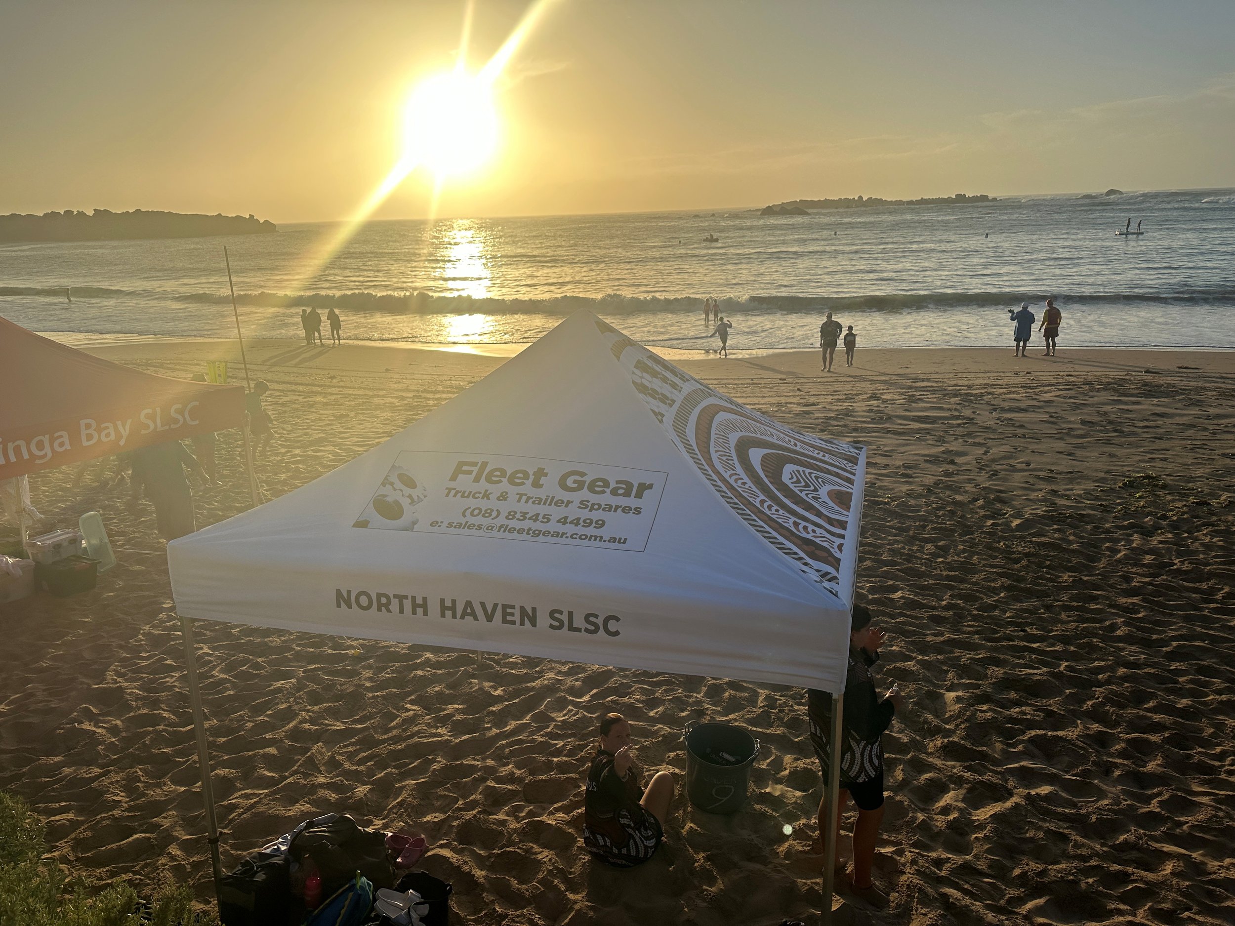 Sunset on a beach with people walking by the water, a white canopy tent labeled 'North Haven SLSC' and 'Fleet Gear Truck & Trailer Spares', and a few individuals near the tent.