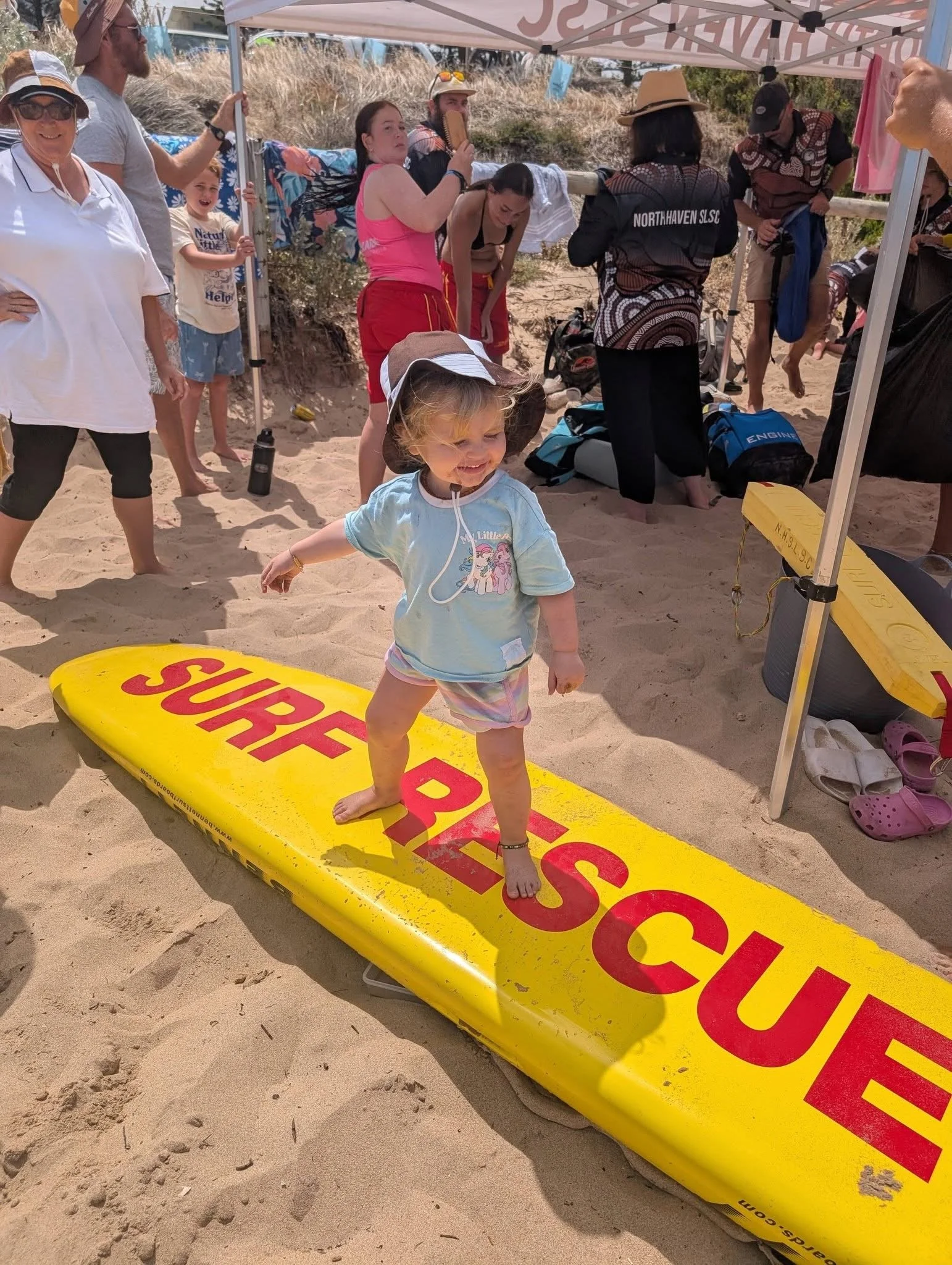 A young girl smiling while standing barefoot on a yellow surfboard with red text on a sandy beach. She is wearing a light blue shirt, colorful shorts, a sun hat, and an ankle bracelet. Several people are gathered behind her under a tent, some looking