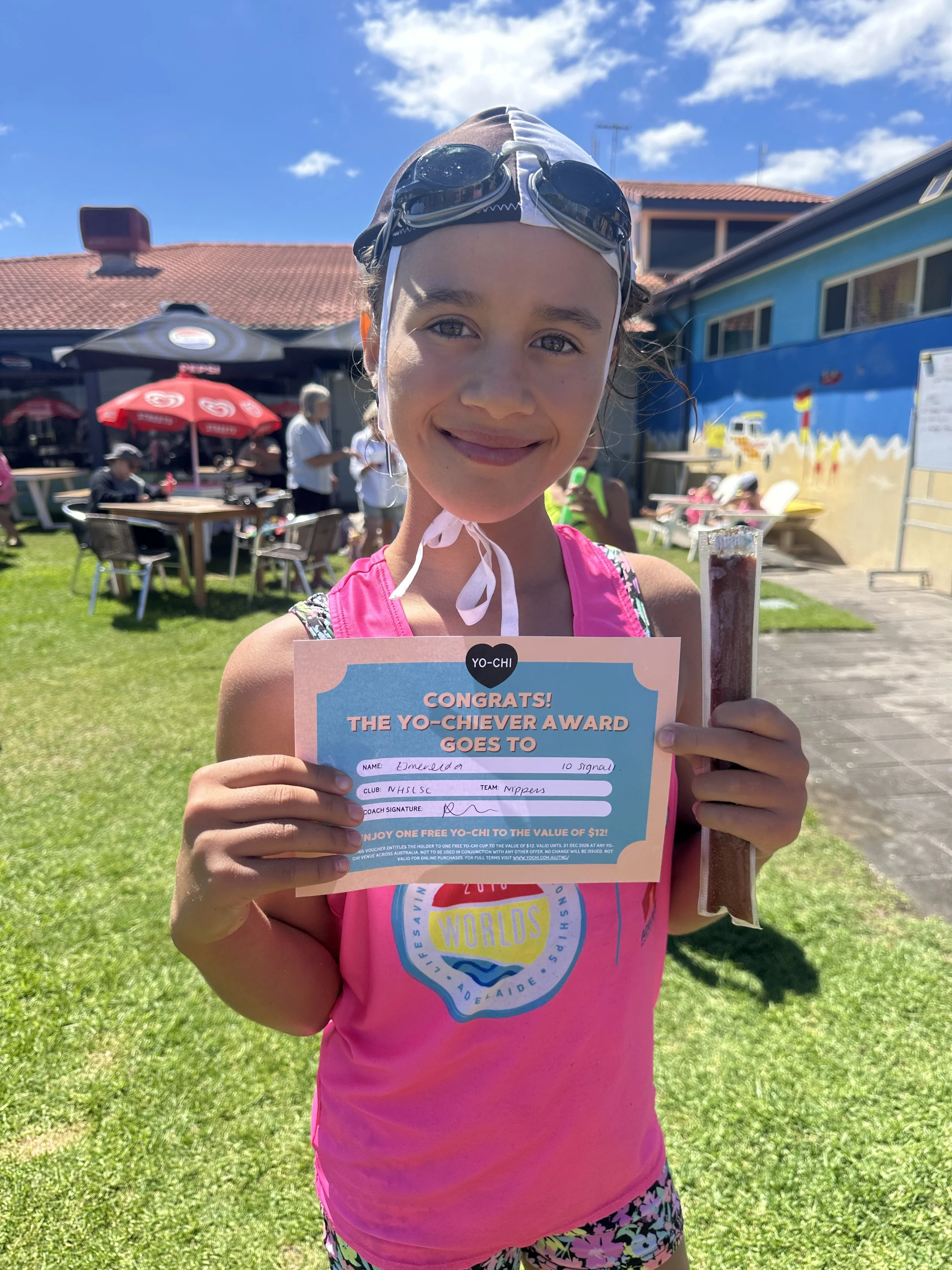 A young girl in a pink tank top holding a certificate and a popsicle outdoors on a sunny day. She is wearing swimming goggles and smiling.