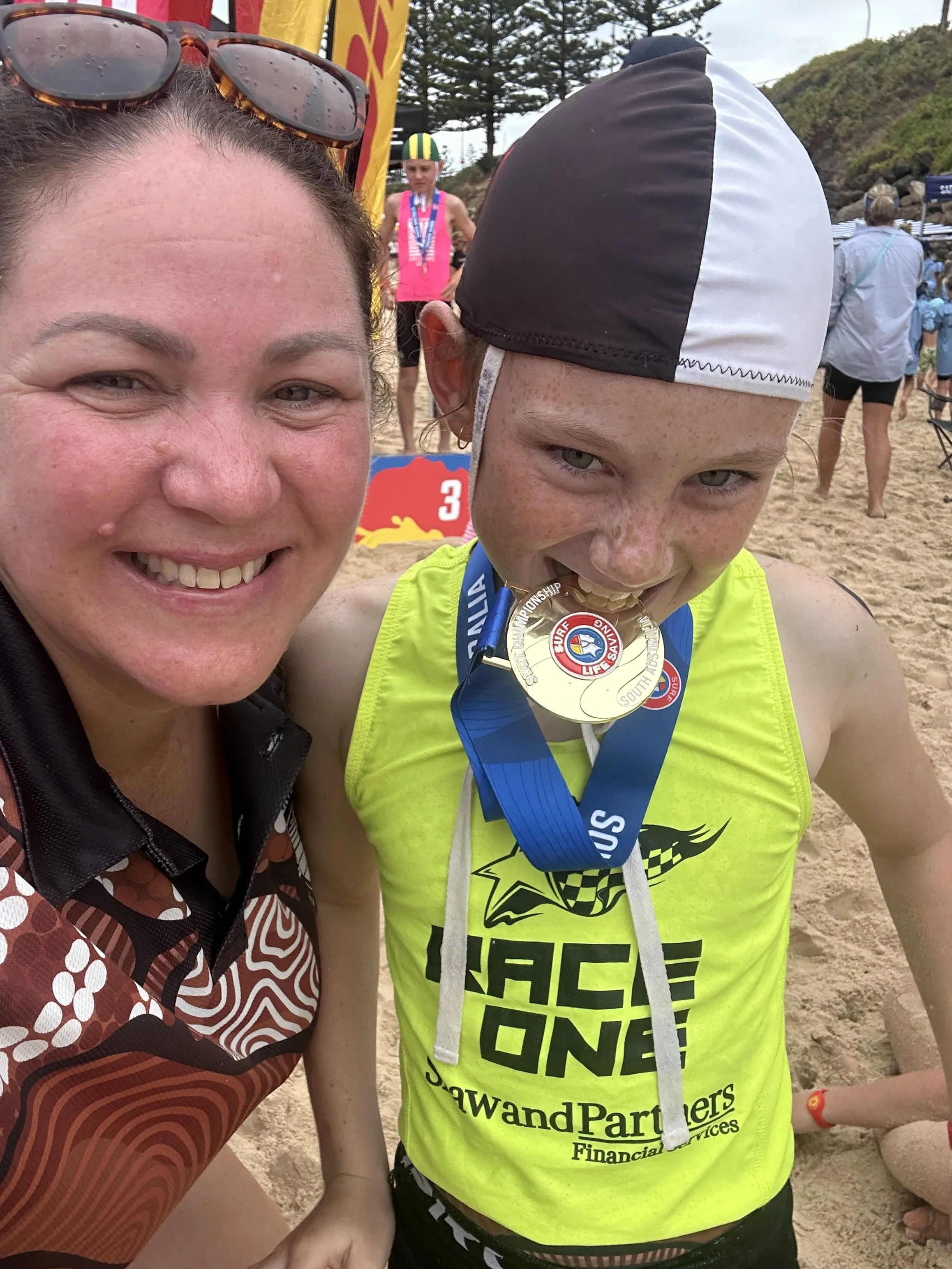 A woman and a young girl at the beach. The girl, wearing a yellow sports shirt, is biting a gold medal at a surfing competition, with other people and tents in the background.