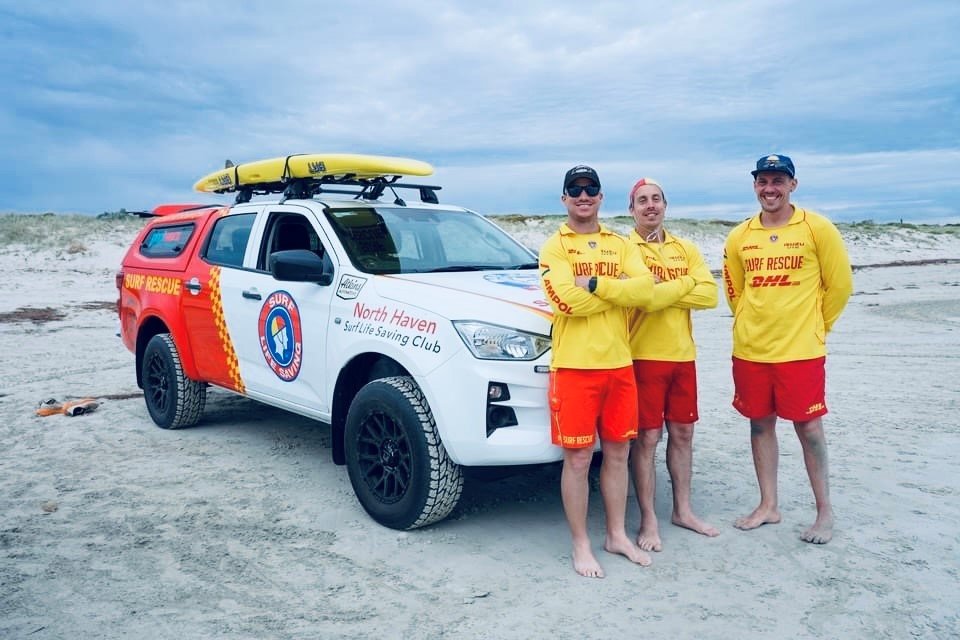 Three surf rescue team members in yellow and red uniforms standing on the beach next to a rescue vehicle with surf rescue logo, surfboards on top, and text 'North Haven Surf Life Saving Club' on the side.