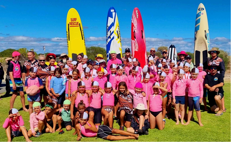 Group of children and adults at the beach, many kids wearing pink shirts and swim caps, standing in front of colorful paddleboards on a grassy area with blue skies overhead.