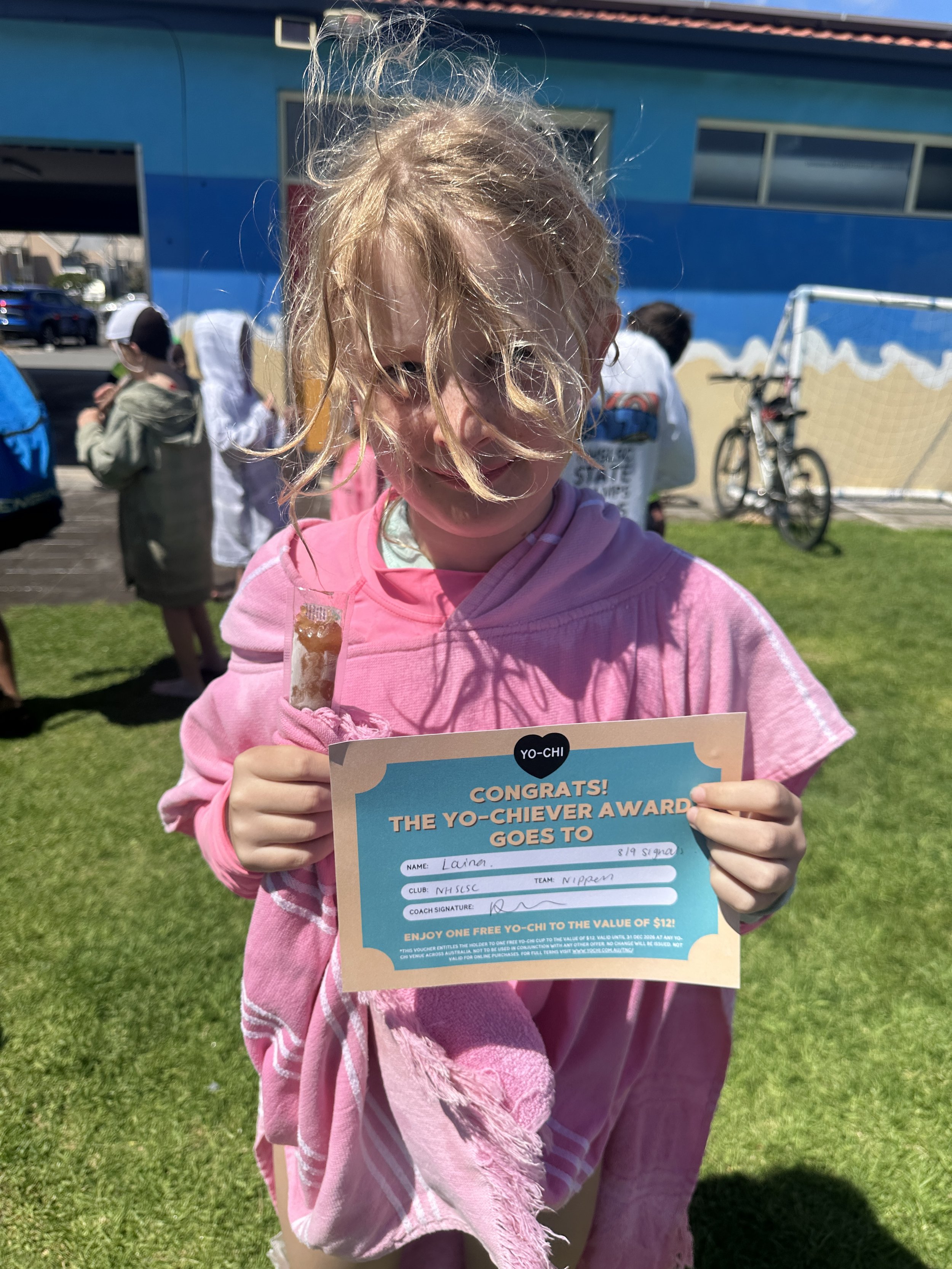 A young girl with curly blonde hair, wearing a pink hoodie, holding a certificate and a marshmallow treat, standing on a grassy field on a sunny day. Several people and a blue building are in the background.