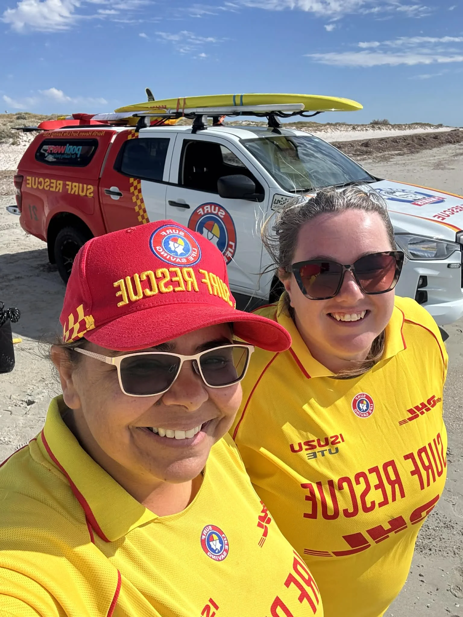 Two women in yellow shirts smiling for a selfie on the beach, with a rescue vehicle and surfboards on top behind them.