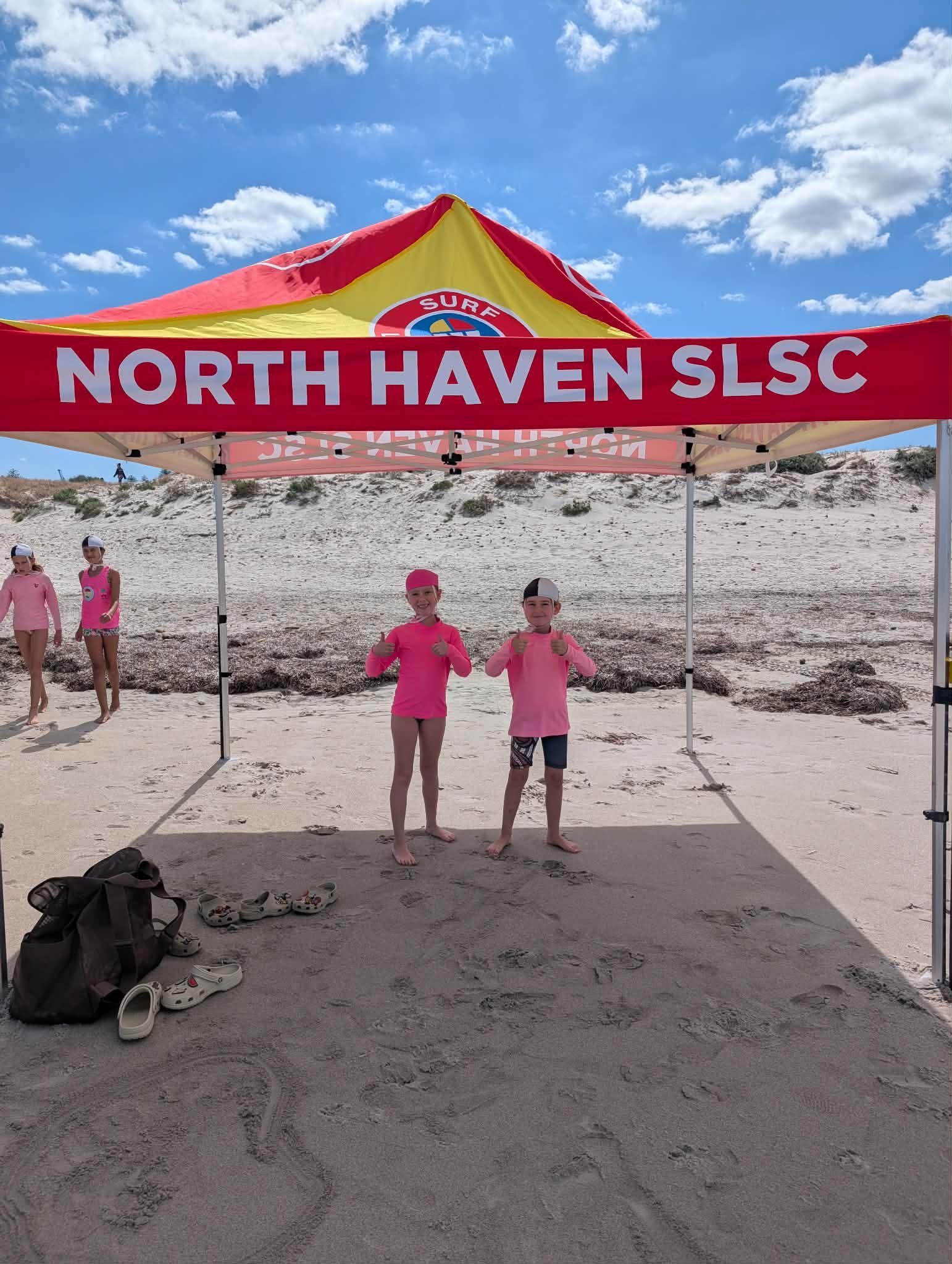 Two children in pink shirts standing under a red and yellow tent on a sandy beach, giving thumbs up, with other children in the background, and a clear blue sky with some clouds.