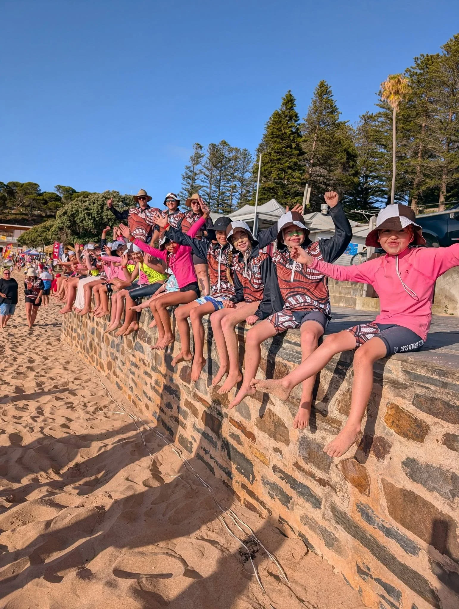 Group of children sitting on a stone wall at the beach, with some wearing hats and relaxing under the sun.