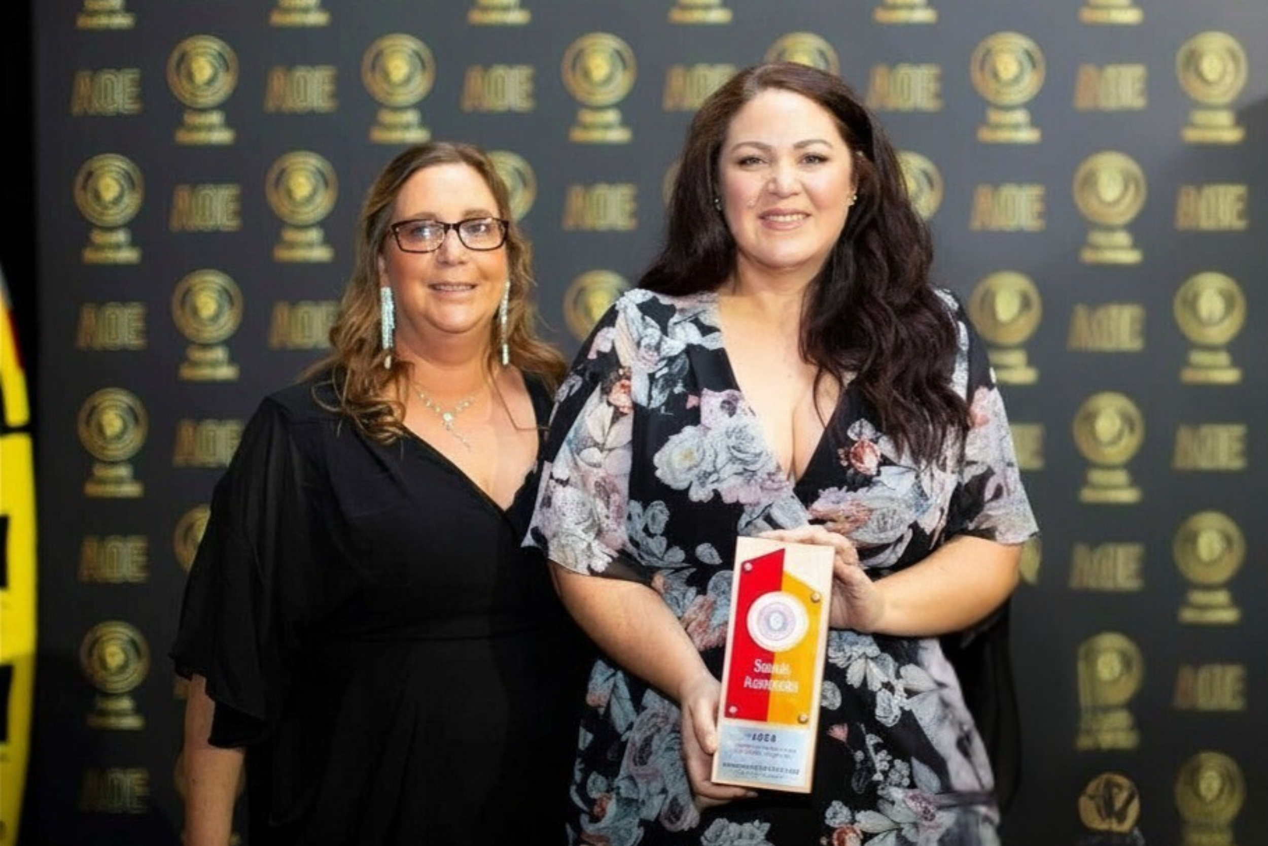 Two women standing together at an award ceremony, one holding a trophy or award, with a backdrop featuring a circular emblem and the word 'MOE' repeatedly printed.