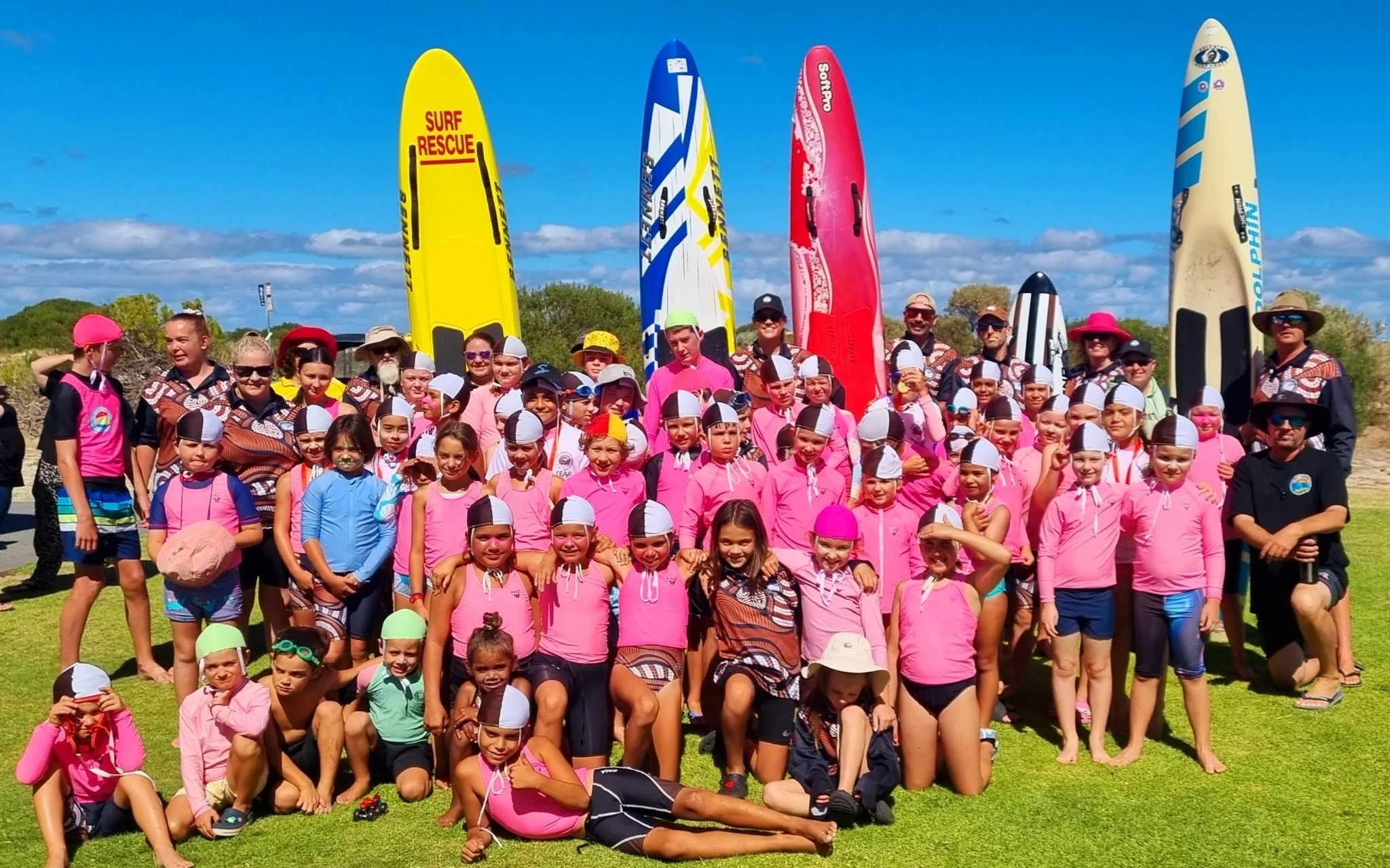 Group of children and adults gathered outdoors on a sunny day, with several colorful surfboards standing upright behind them.
