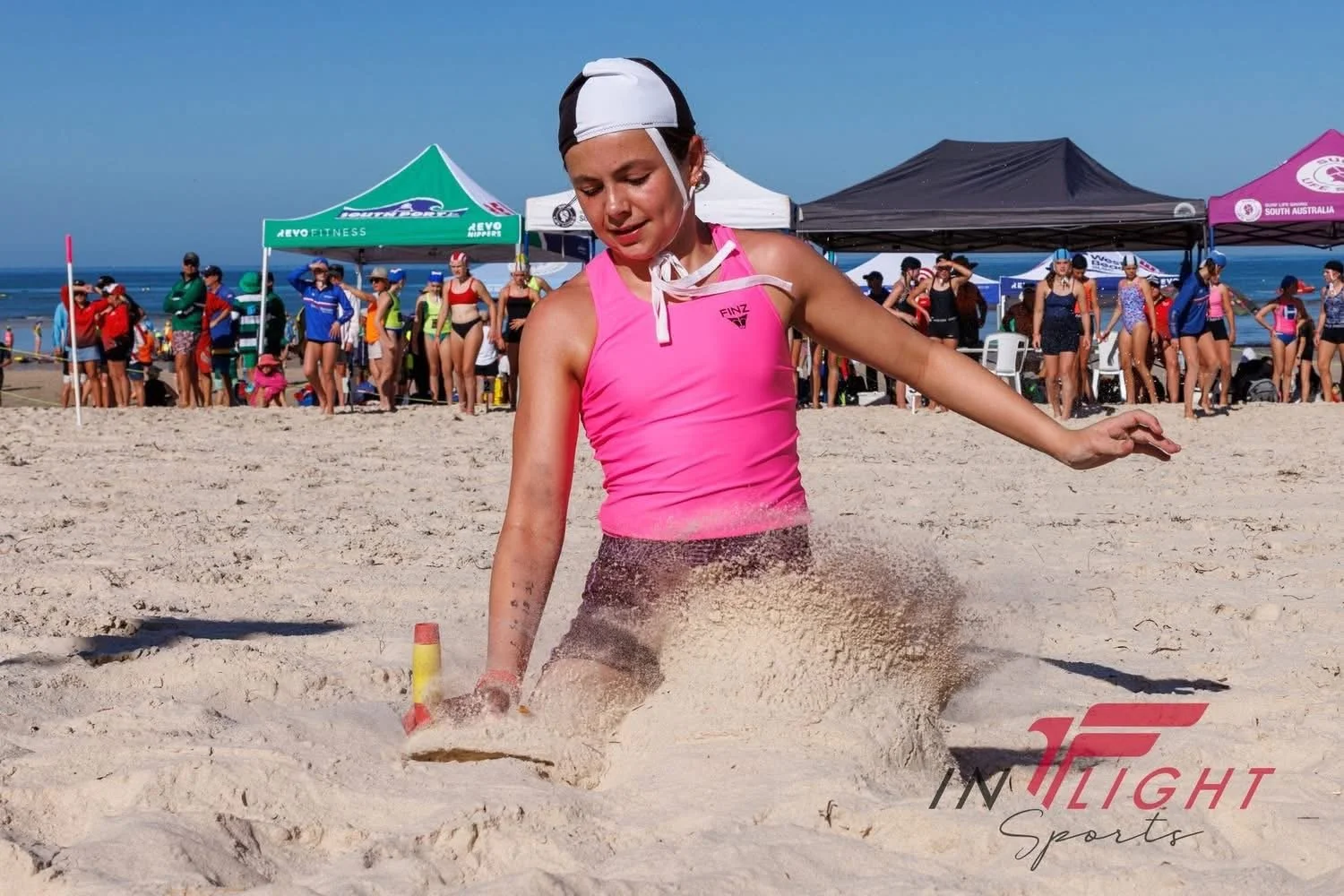 A young girl in a pink sports top and black and white swimming cap is sitting in the sand during a beach event, with a large group of people and several tents in the background, and the ocean visible beyond.