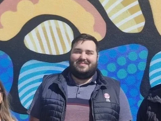 A man with a beard standing in front of a colorful abstract mural.