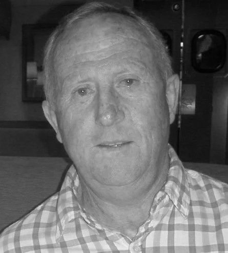 Black and white photo of a middle-aged man with light skin, short hair, wearing a checkered button-up shirt, smiling slightly.