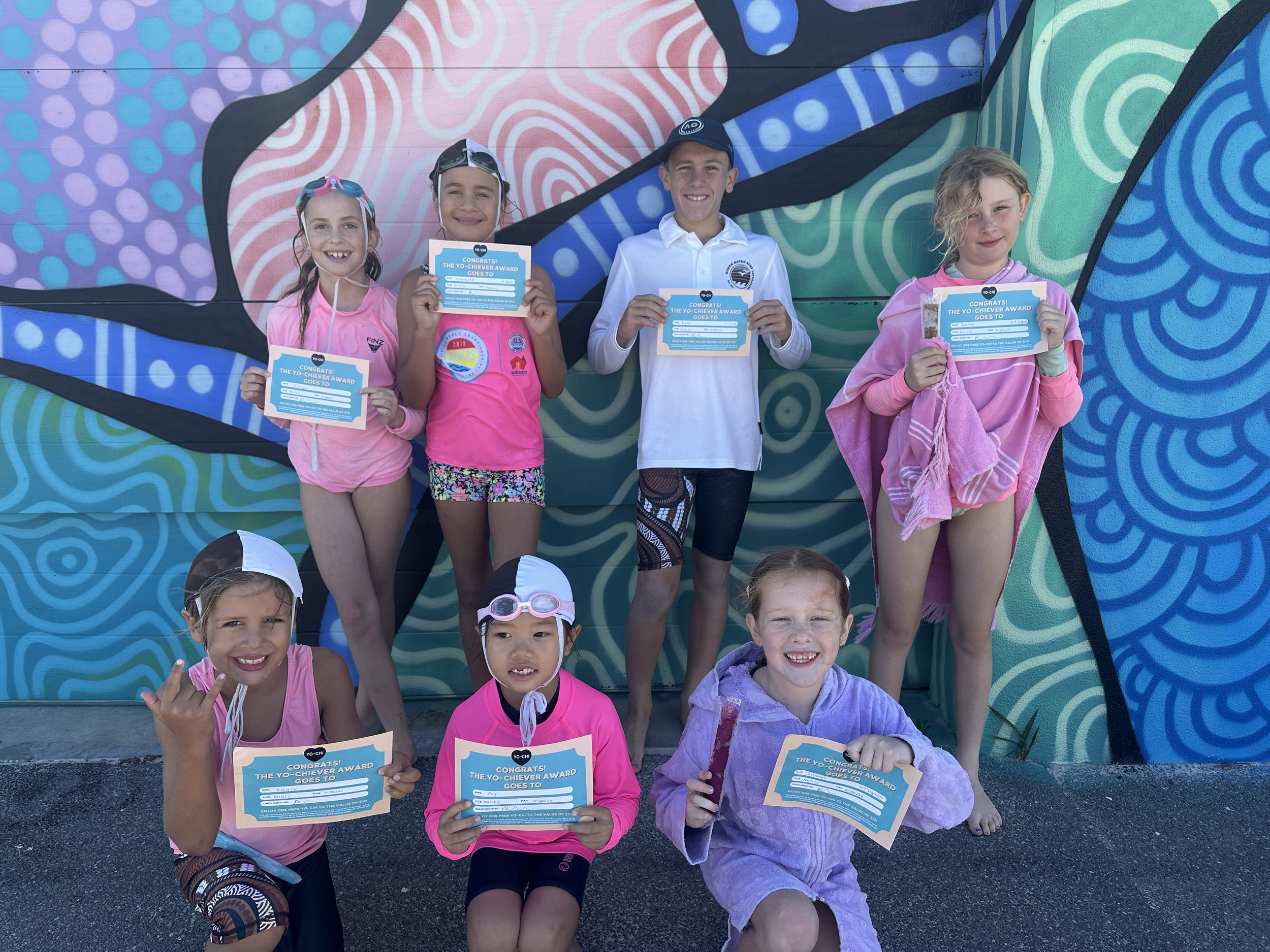 Seven children, four girls and three boys, holding blue certificates with the text "Congratulations! The Yo-Chieve Award Goes To" in front of a colorful, abstract mural.