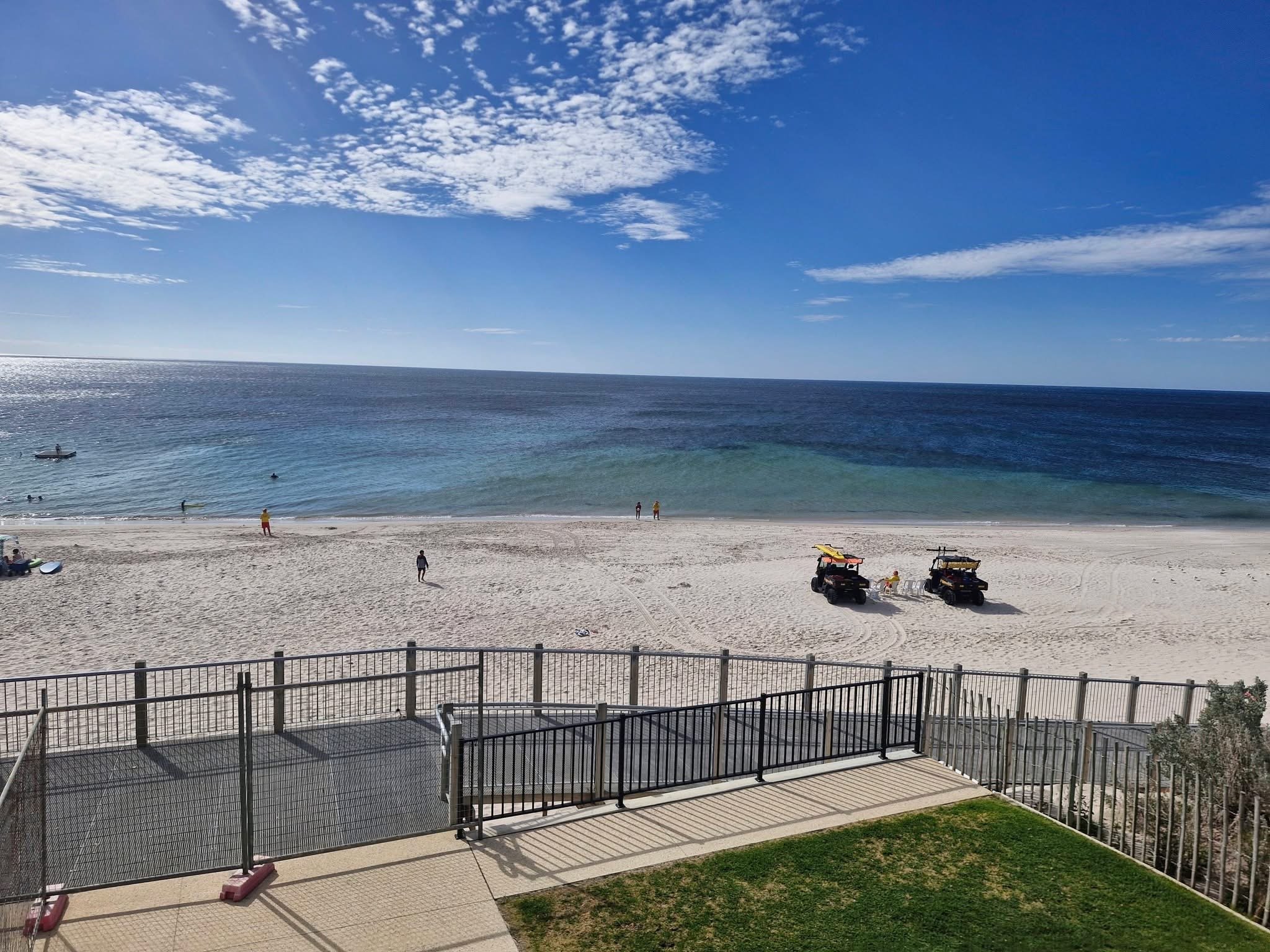 View of a sandy beach with a few people, two off-road vehicles, and a body of water with gentle waves under a partly cloudy sky, seen from a balcony with a black metal fence.