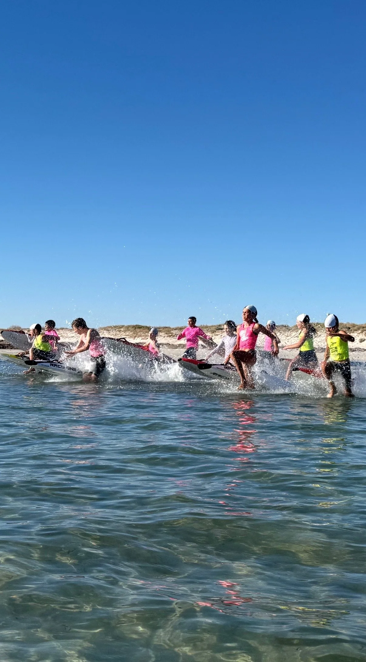 Group of children in life jackets learning to stand and paddle on surfboards on the beach under a clear blue sky.