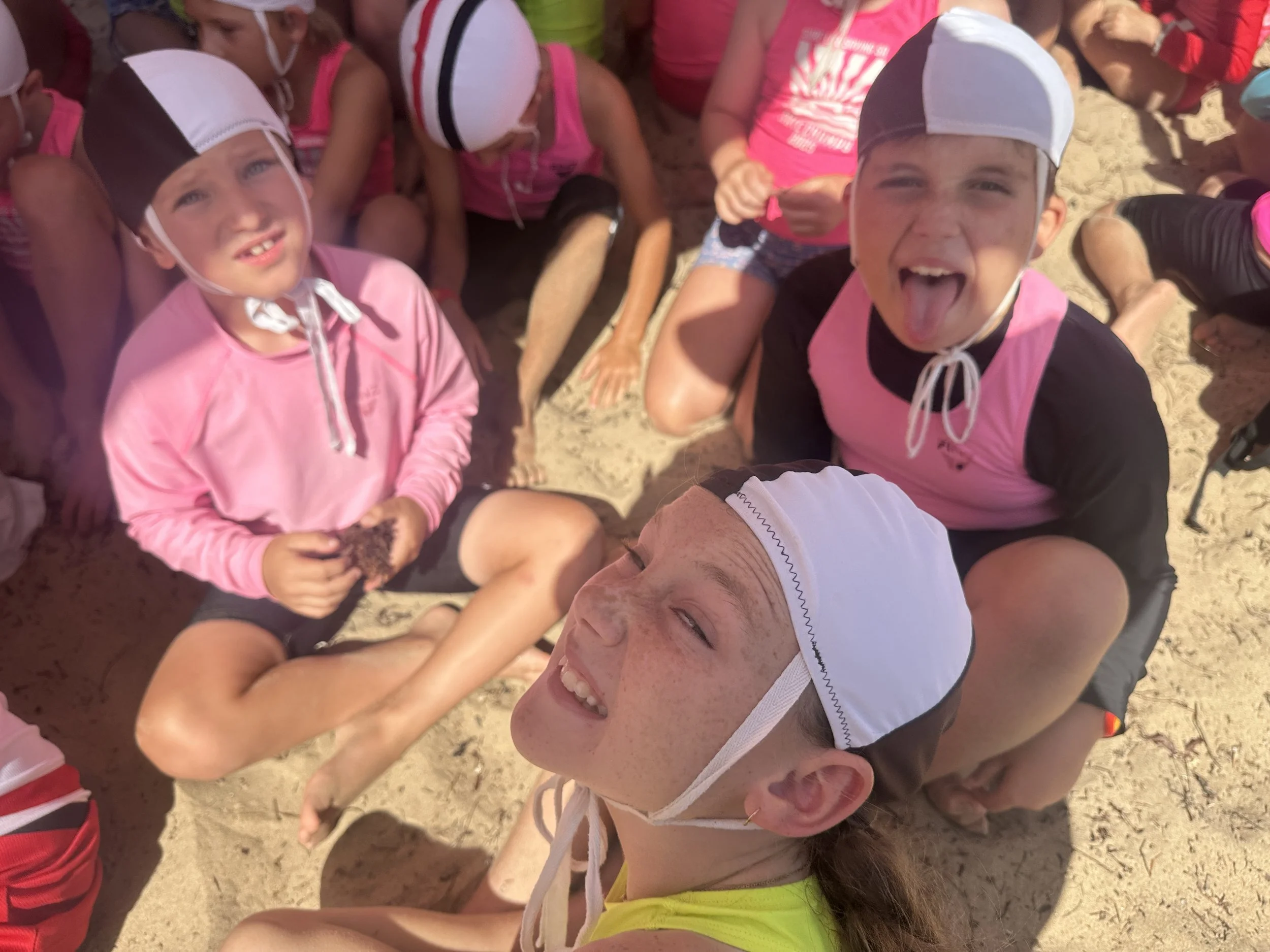 Children wearing swim caps and pink tops are sitting on the sand at the beach, enjoying a sunny day.