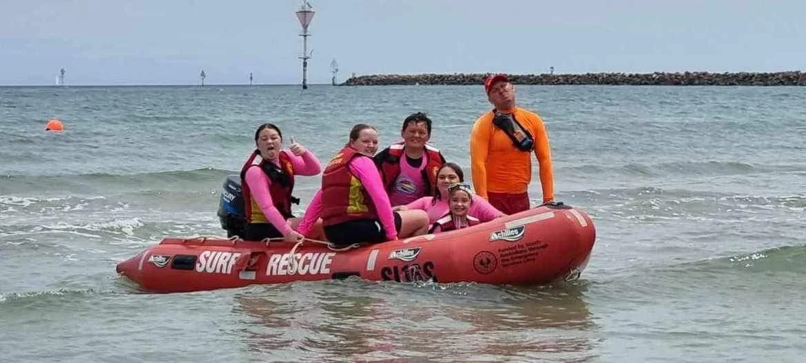 Group of people sitting and standing on a red rescue boat in the ocean with a breakwater and navigation markers in the background.