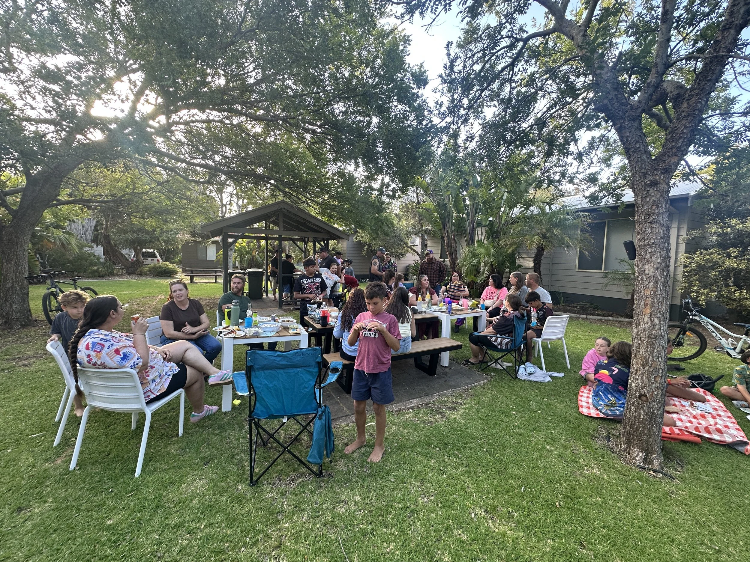 Group of people gathered for an outdoor celebration or party in a backyard, with tables, chairs, and trees.