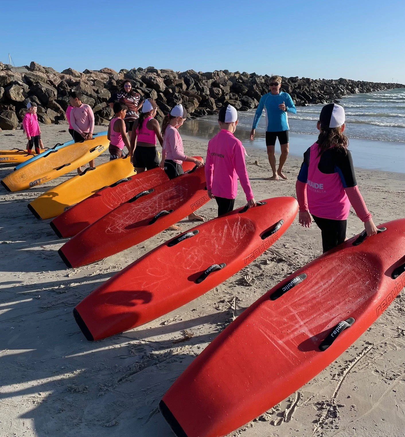 Group of children in pink and black wetsuits with skull caps on a sandy beach, getting ready for a board paddling lesson with two instructors, one in a blue shirt and one in a patterned shirt, near boards of red and yellow color.