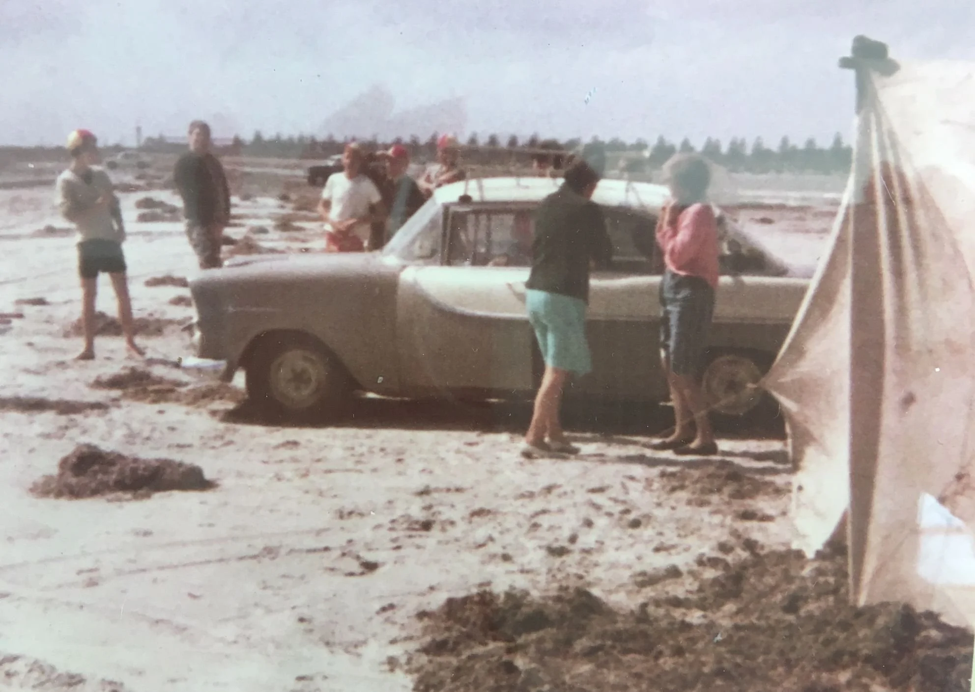 People standing around a vintage car on a muddy field, with some smoking in the background and a large tent on the right side.