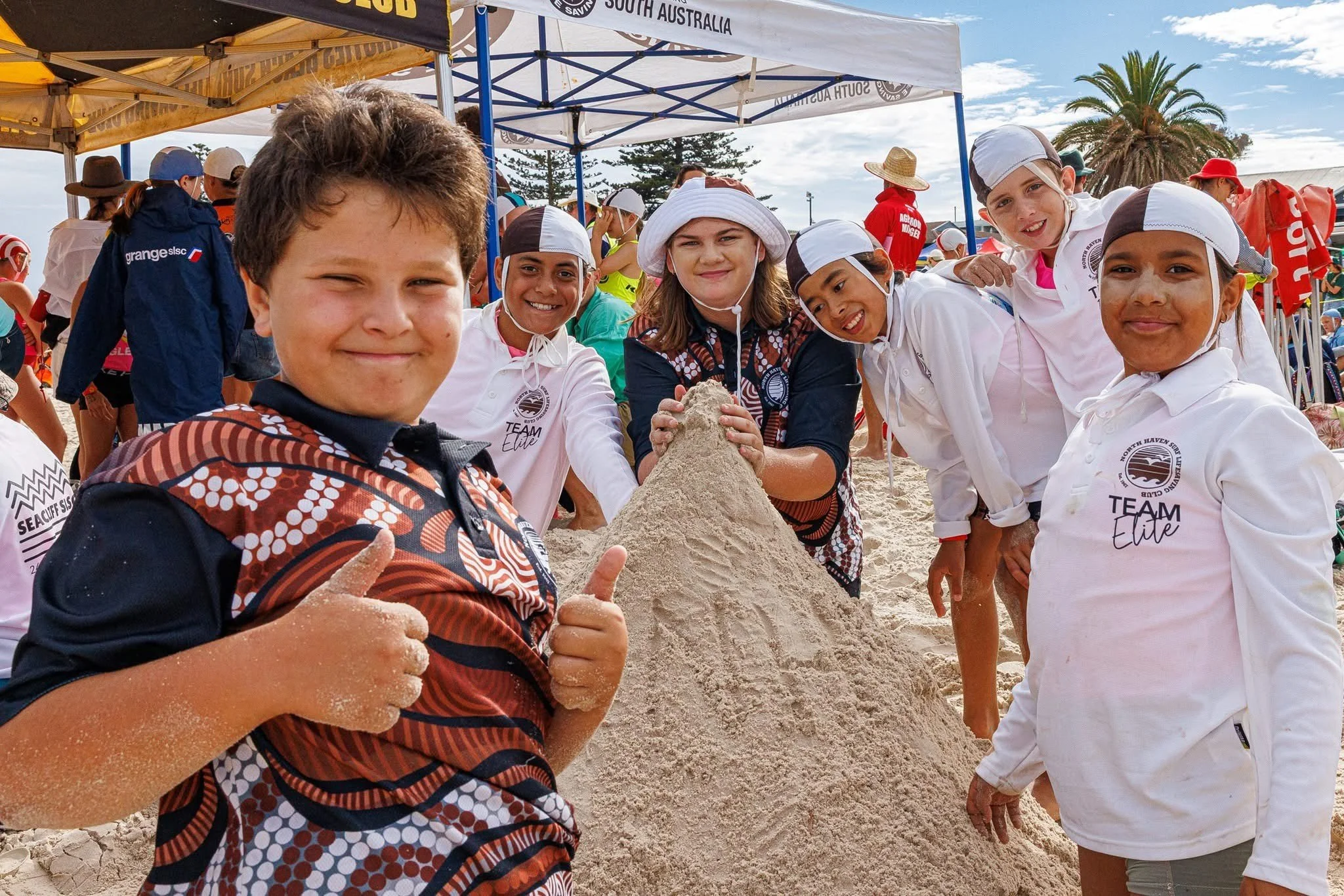 Group of children playing on a sandcastle at a beach event, smiling and giving thumbs-up signs, with tents and palm trees in the background.