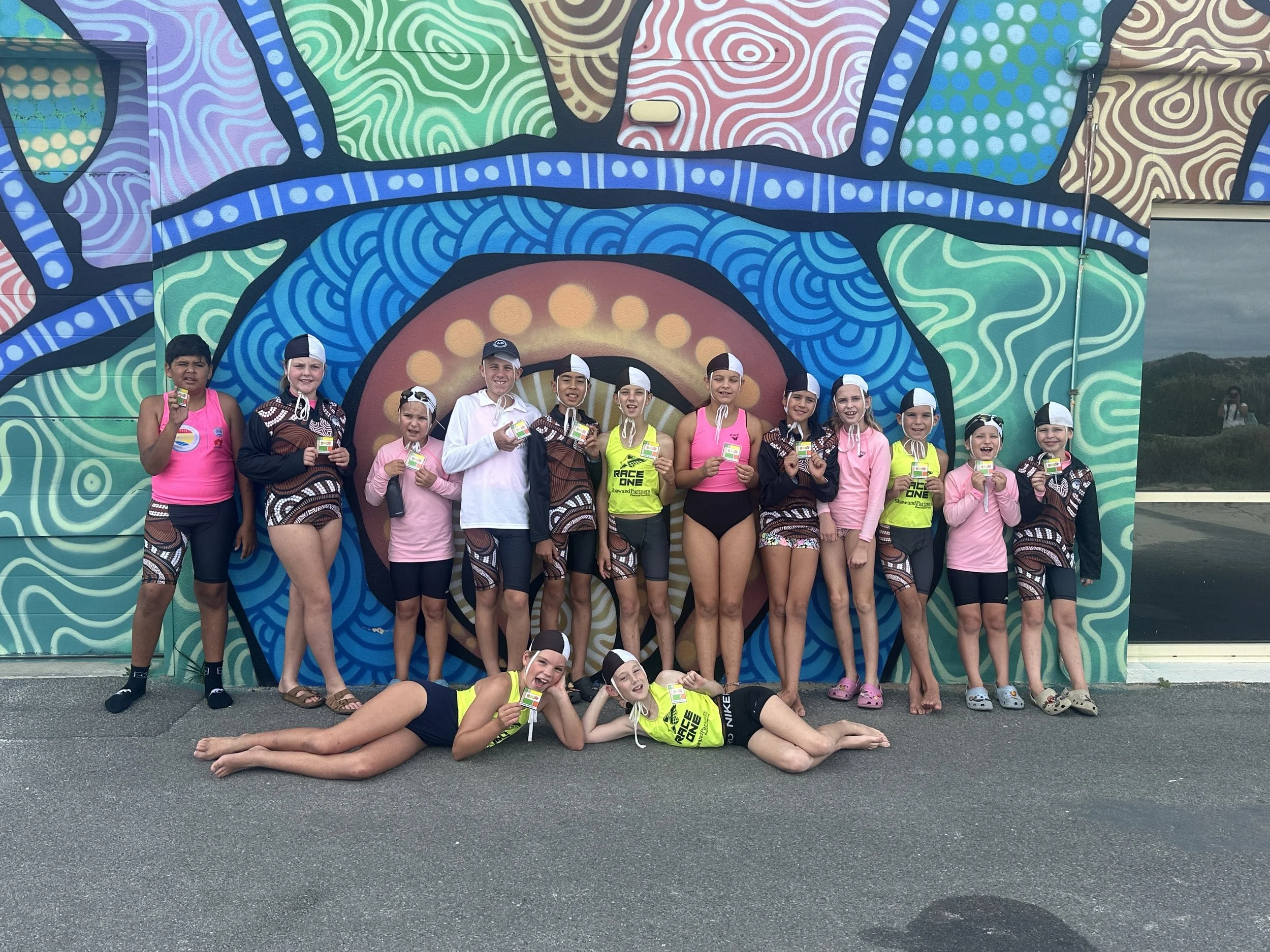 Group of young children standing and lying in front of a colorful, abstract mural, wearing swimwear and holding medals, with some children in pink shirts and others in matching brown and white patterned swimwear.
