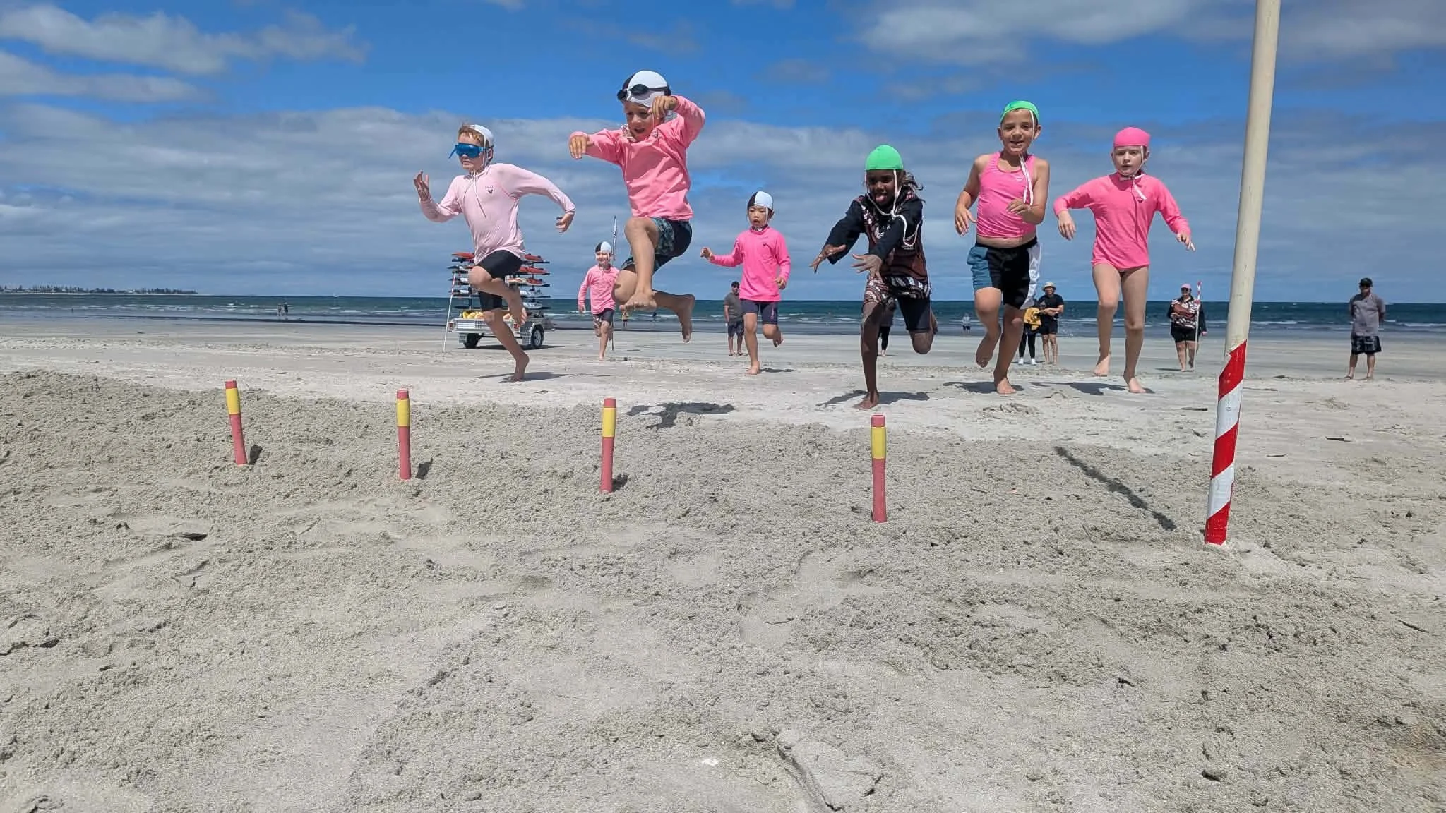 Children on a beach jumping over a sand obstacle course with flags, under a partly cloudy sky.