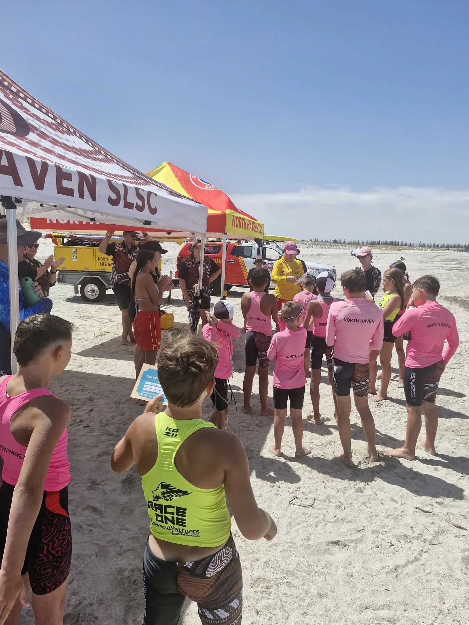 Children and adults gathered on a sandy beach for a rescue instruction demonstration, with tents and rescue vehicles in the background.