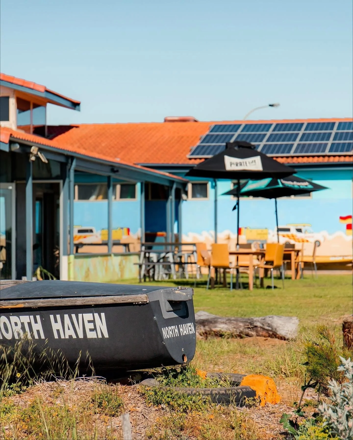 A boat with the words 'North Haven' on it, on grass with plants in the foreground. In the background, there is a light blue building with a mural, tables and chairs, a black umbrella, and solar panels on the red-tiled roof.