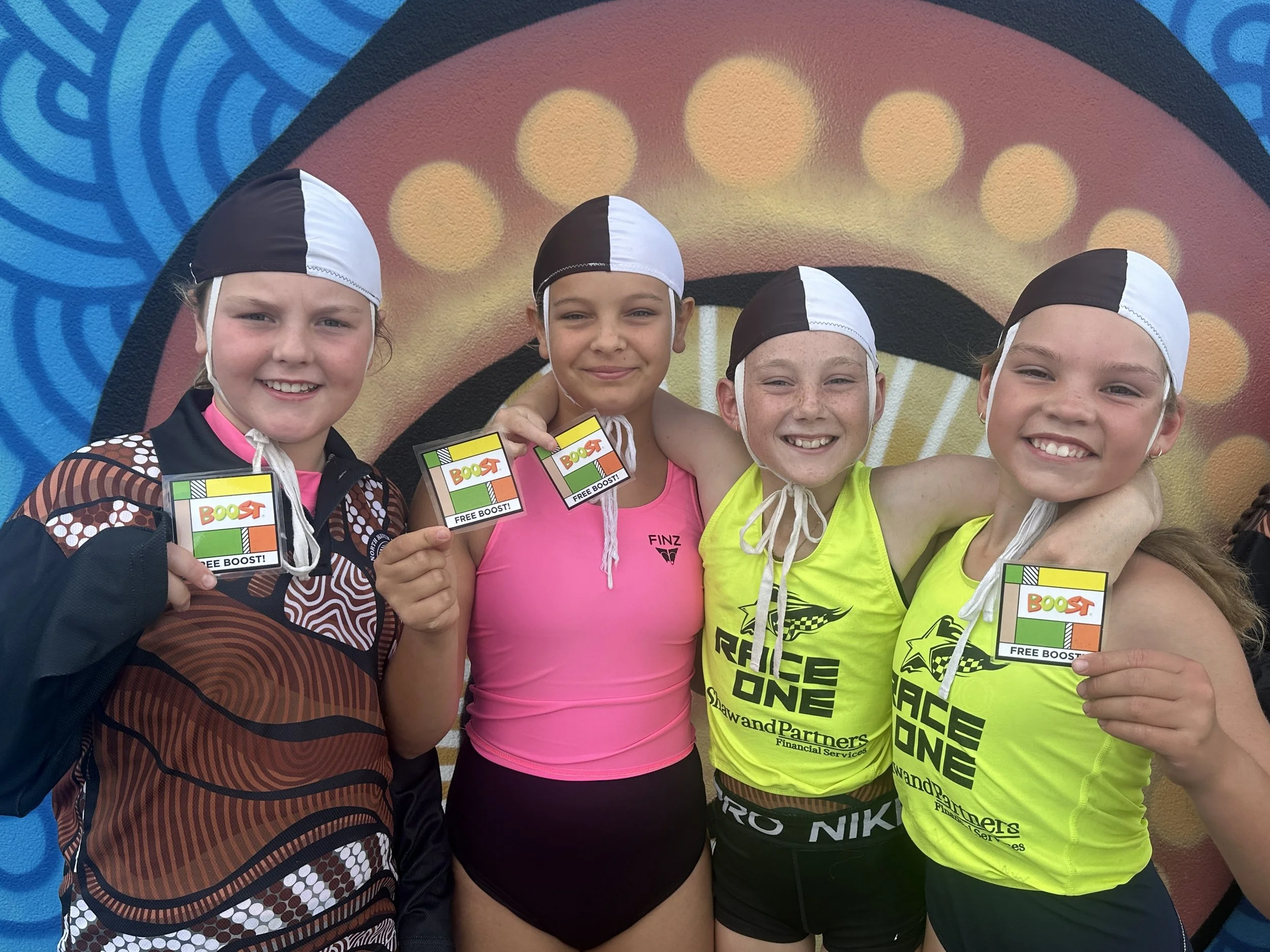 Four young girls in sports attire holding Boost energy drink coupons, smiling in front of a colorful mural.