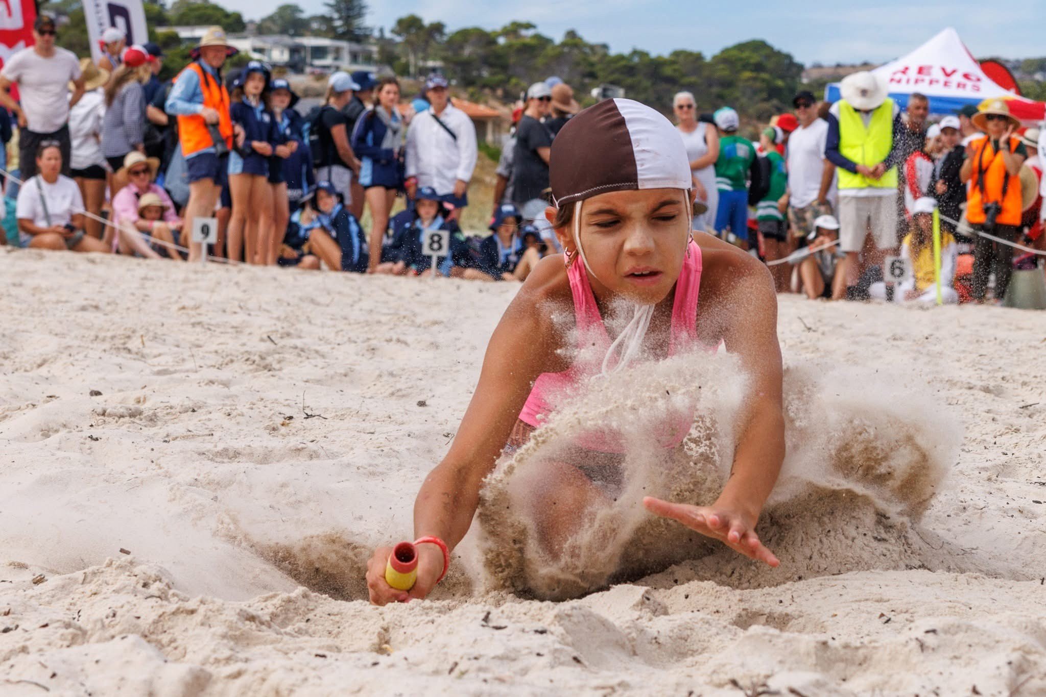 Young girl in pink swimsuit and swimming cap falling in sand during beach event, with crowd of spectators in the background.