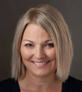 A woman with shoulder-length blonde hair smiling against a dark background.