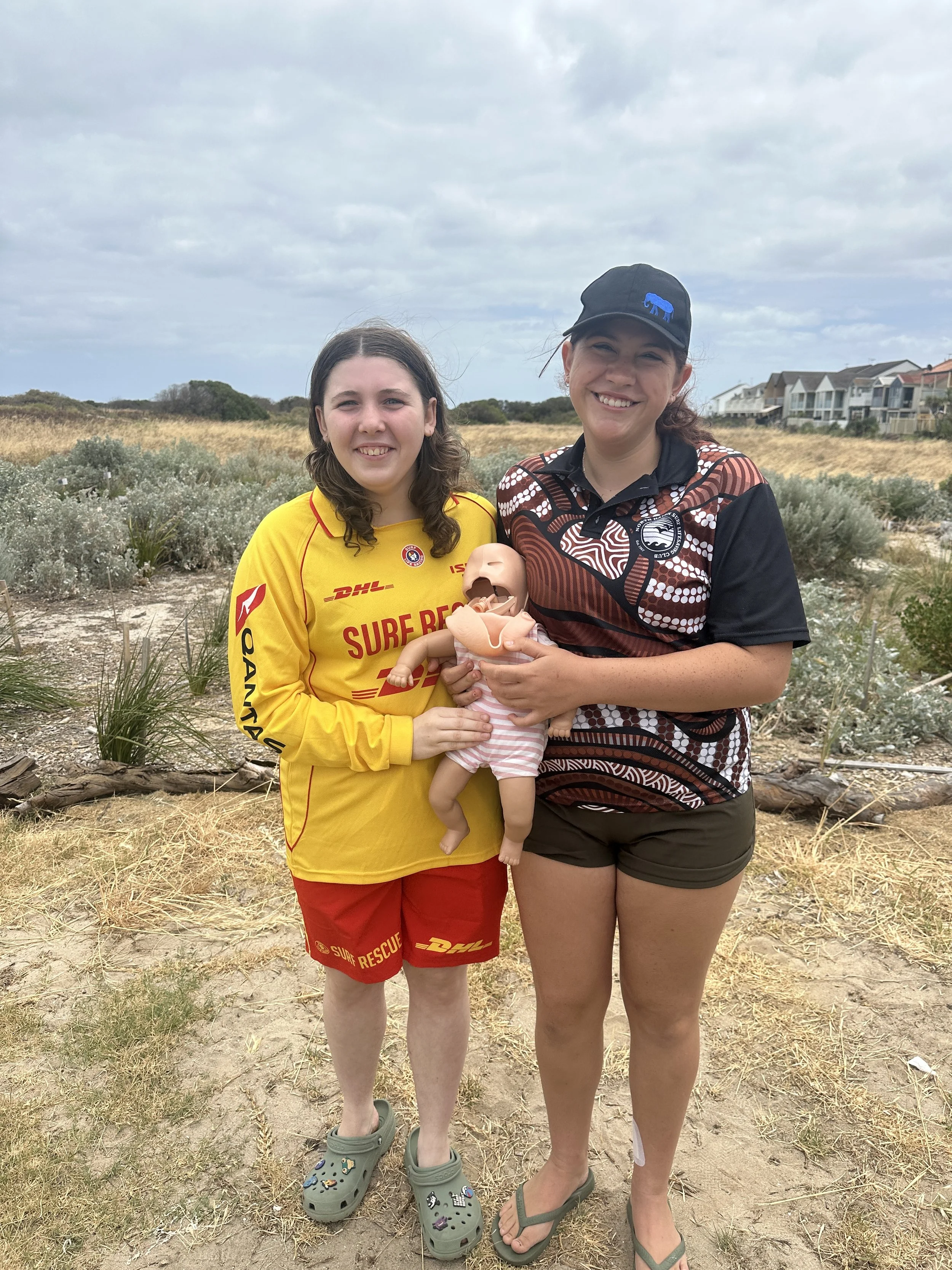 Two smiling women standing outdoors on a cloudy day, holding a baby doll, with bushes and houses in the background.