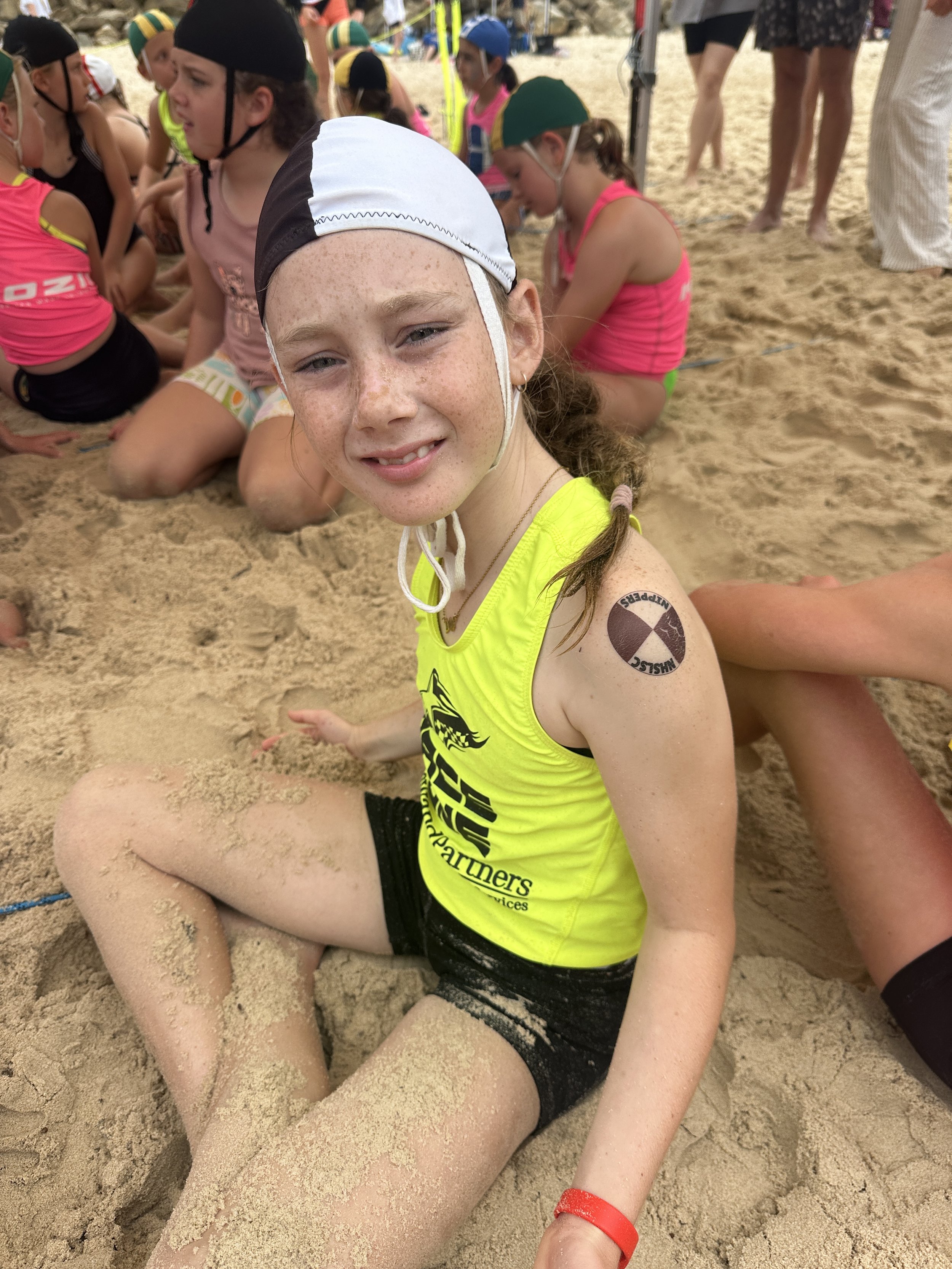 Young girl with a freckled face sitting on sandy ground at a beach or sand sports event, wearing a yellow tank top, black shorts, and a helmet, with other children in the background engaged in activity.