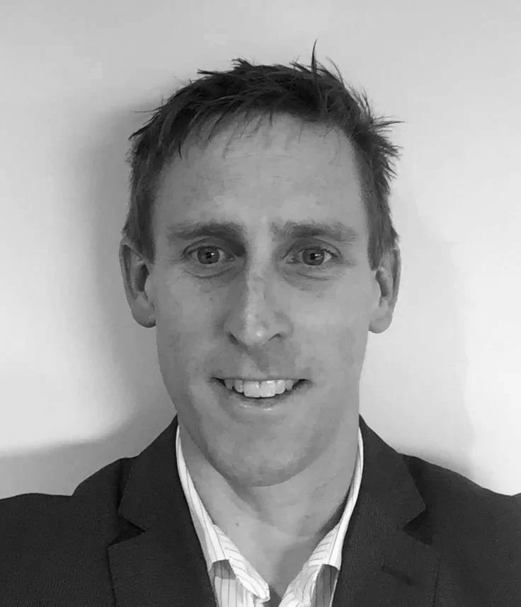 A man with short hair wearing a suit and white shirt, smiling at the camera, in black and white.