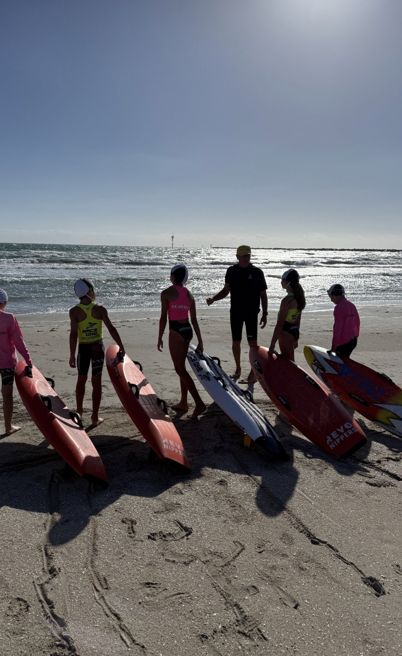 Group of young women with paddleboards and a coach on a sandy beach with the ocean and a lighthouse in the background.