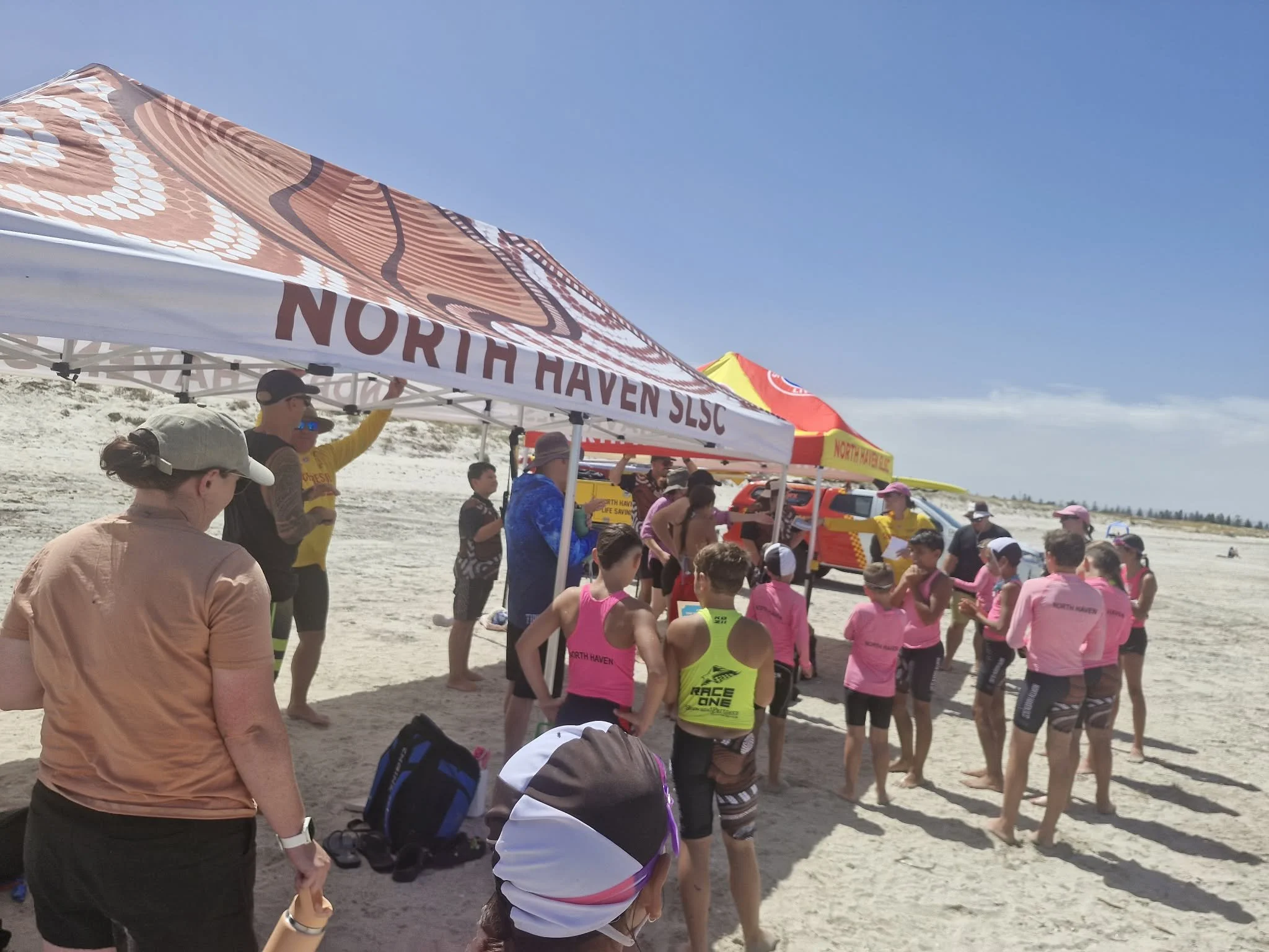 A group of people, including children, gathered under tents on a sandy beach, some wearing running gear and pink shirts, near a rescue vehicle, during a sunny day.