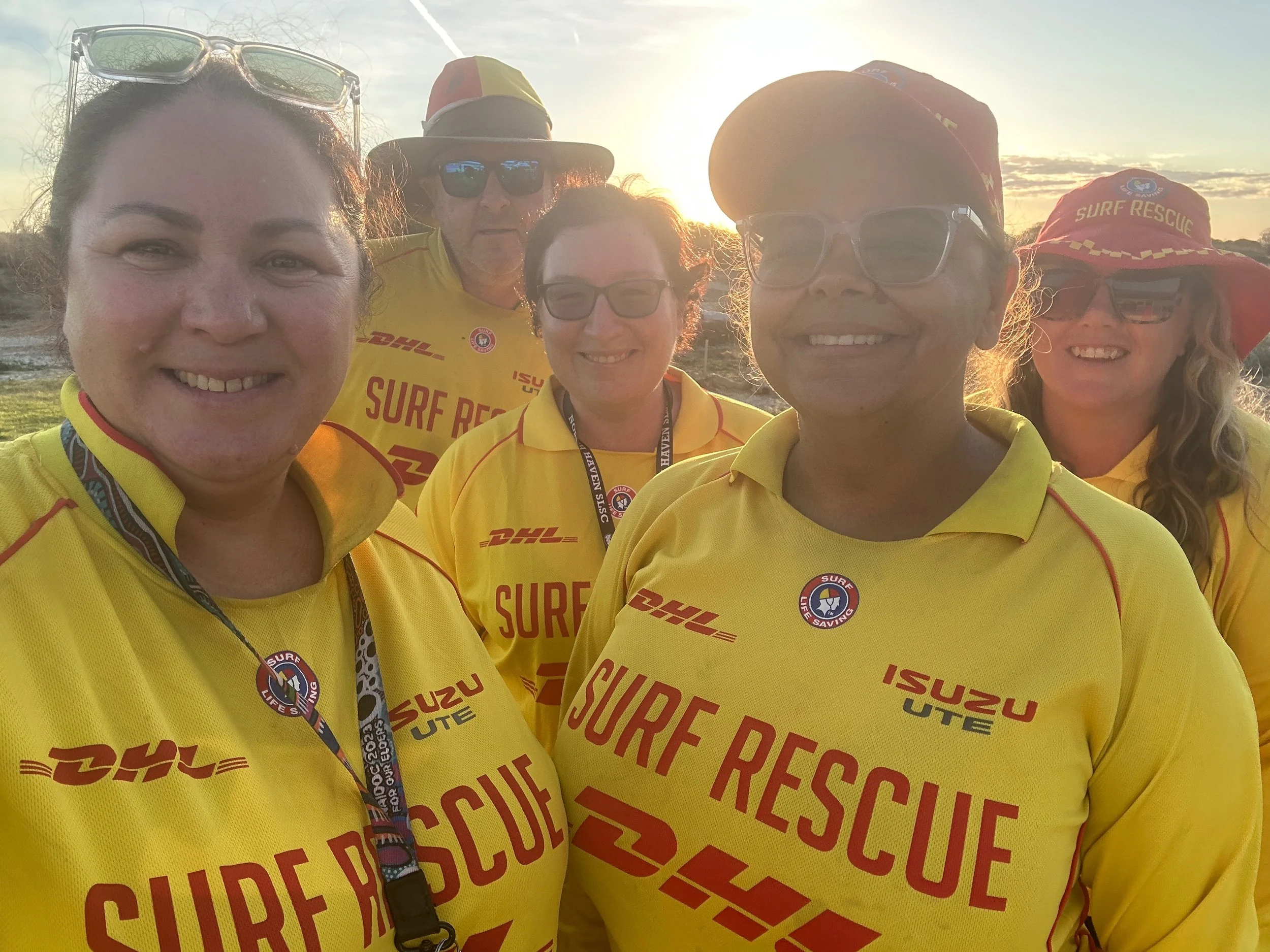 Group of five people in yellow surf rescue shirts taking a selfie outdoors at sunset