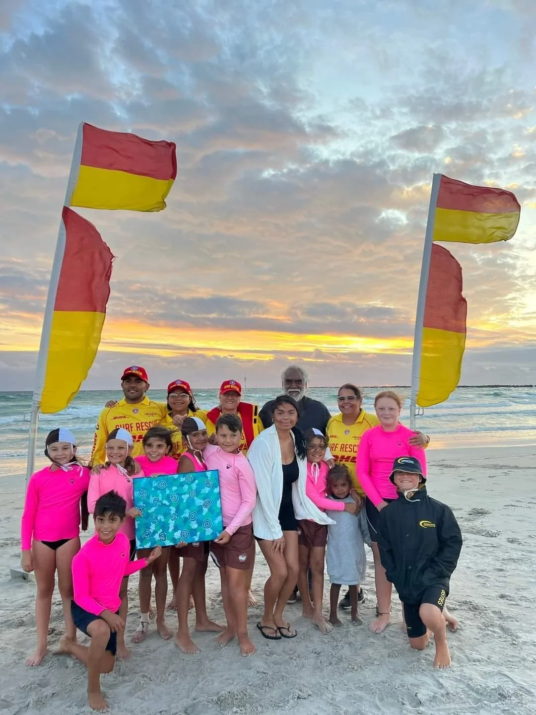 Group of children and adults on a beach at sunset, with flags and rescue personnel in yellow uniforms.
