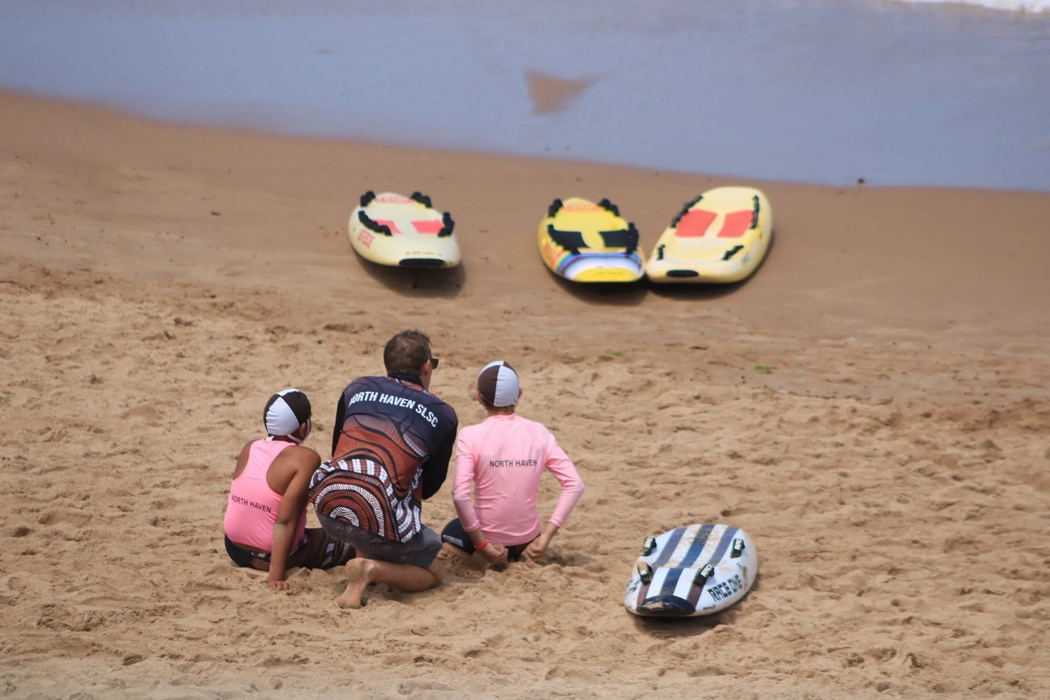 A man and two children sitting on the sandy beach, watching three paddleboards on the sand near the water.