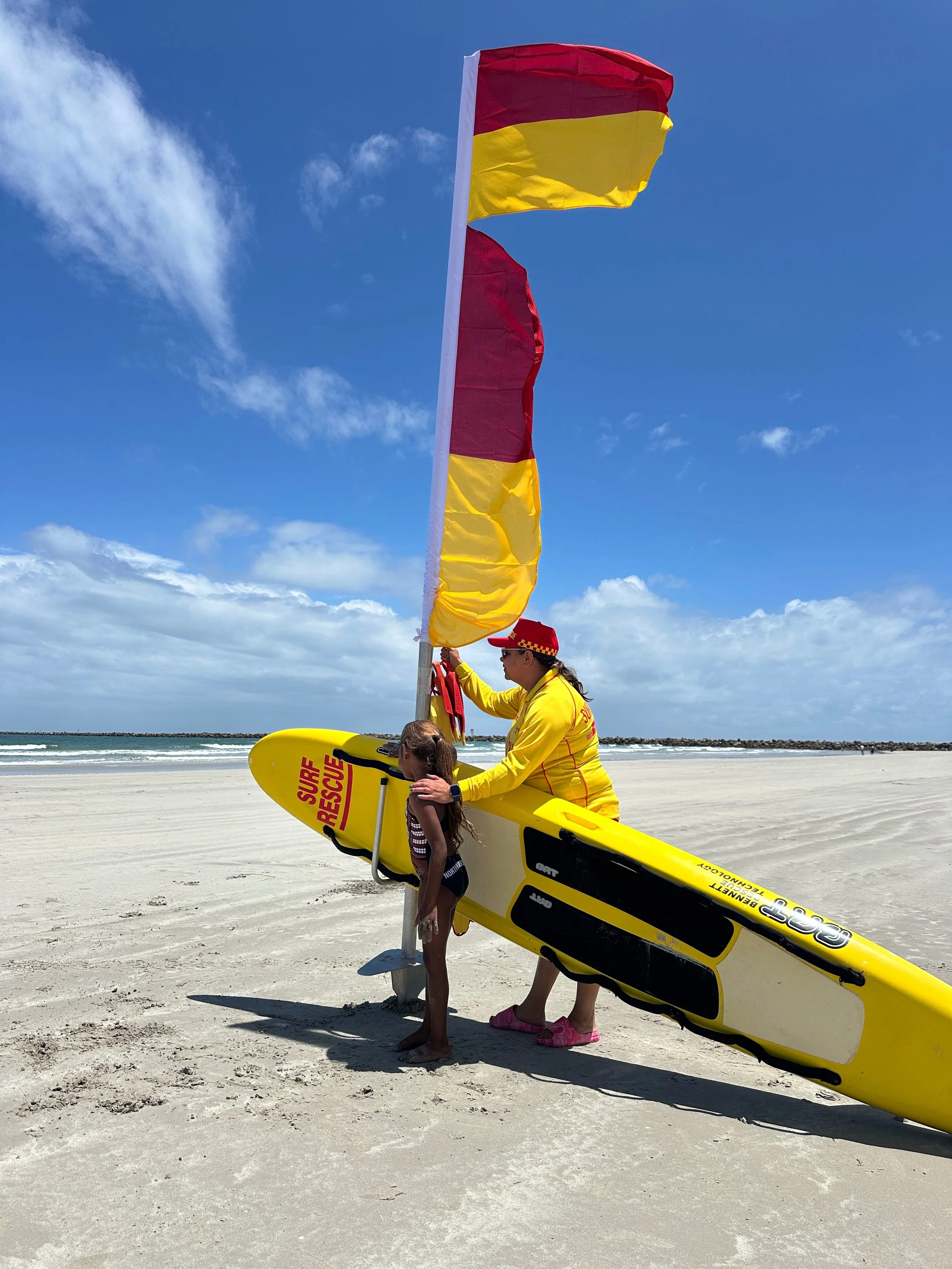 A lifeguard in a yellow uniform holding a bright yellow rescue surfboard, talking to a young girl on a sandy beach. The lifeguard is near a red and yellow flag, with ocean waves and a partly cloudy blue sky in the background.